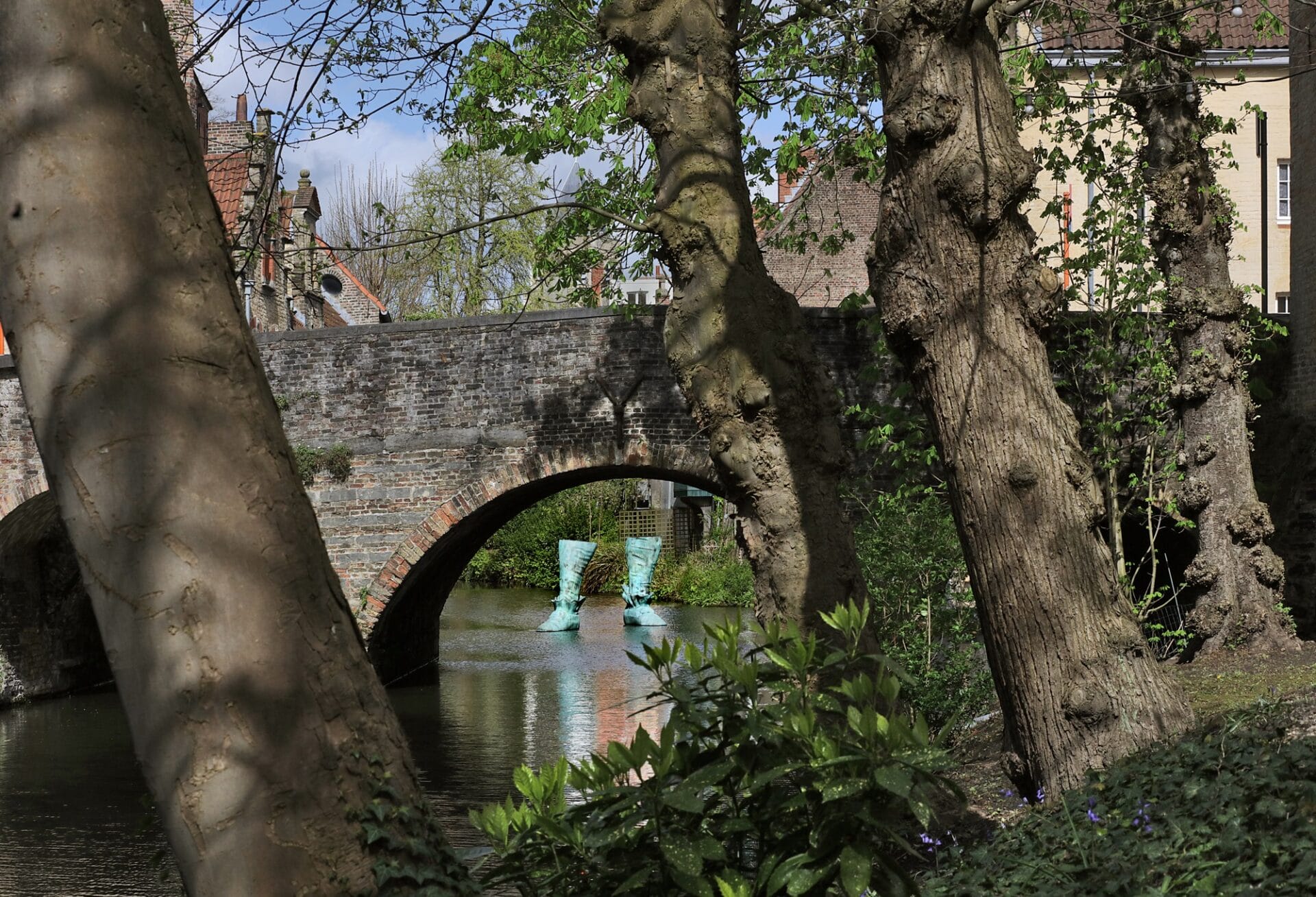 a sculpture of a pair of 18th-century style boots cast in bronze and installed in a bruges canal, seen through some trees and under the arch of a foot bridge