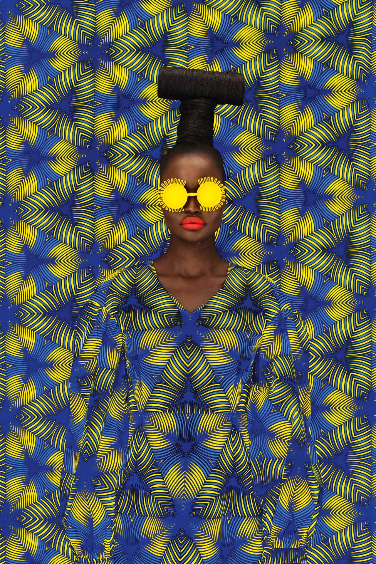 a portrait of the artist against a patterned yellow and blue backdrop. she's wearing bright yellow eyewear