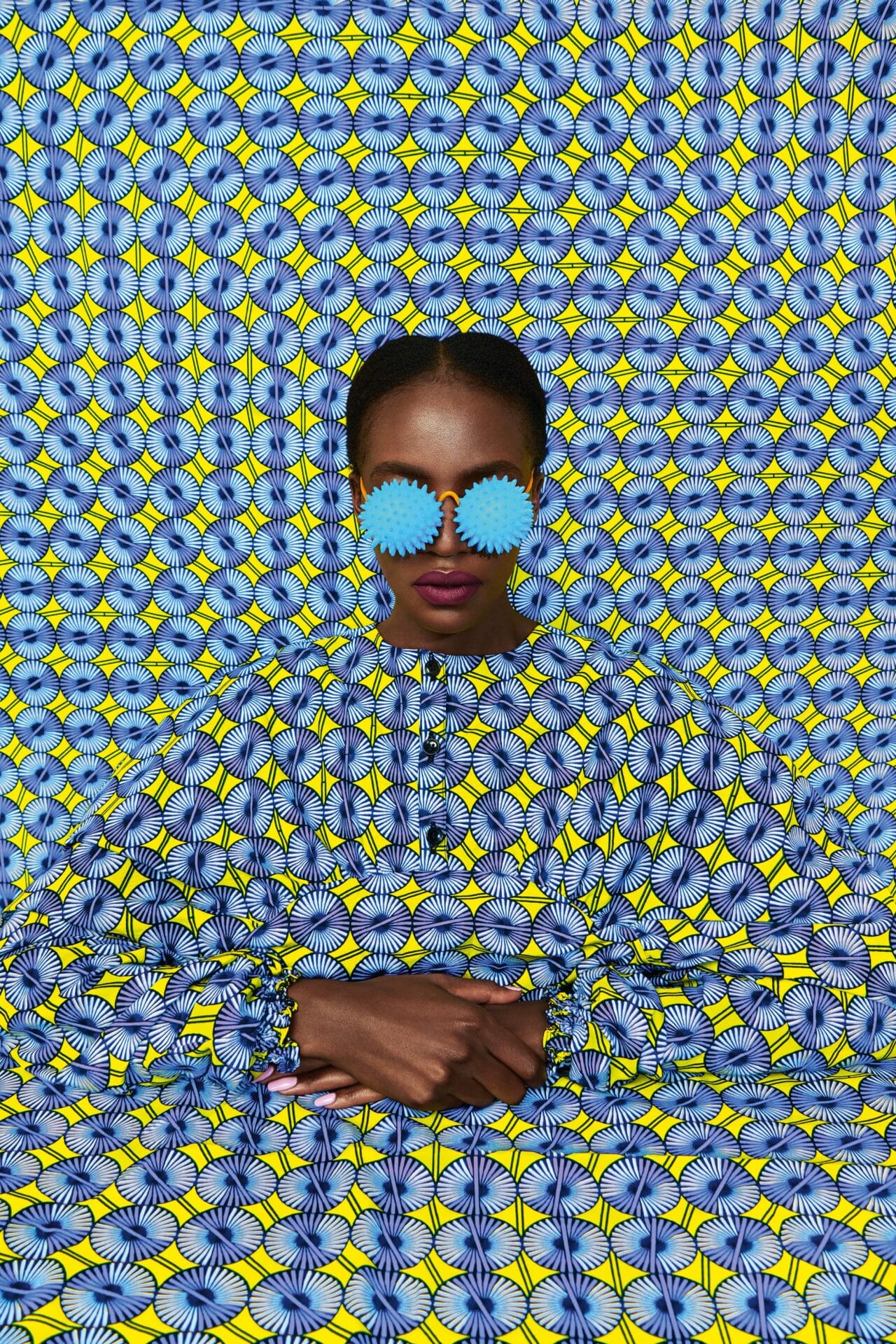 a portrait of the artist against a patterned yellow and blue backdrop. she's wearing light blue eyewear