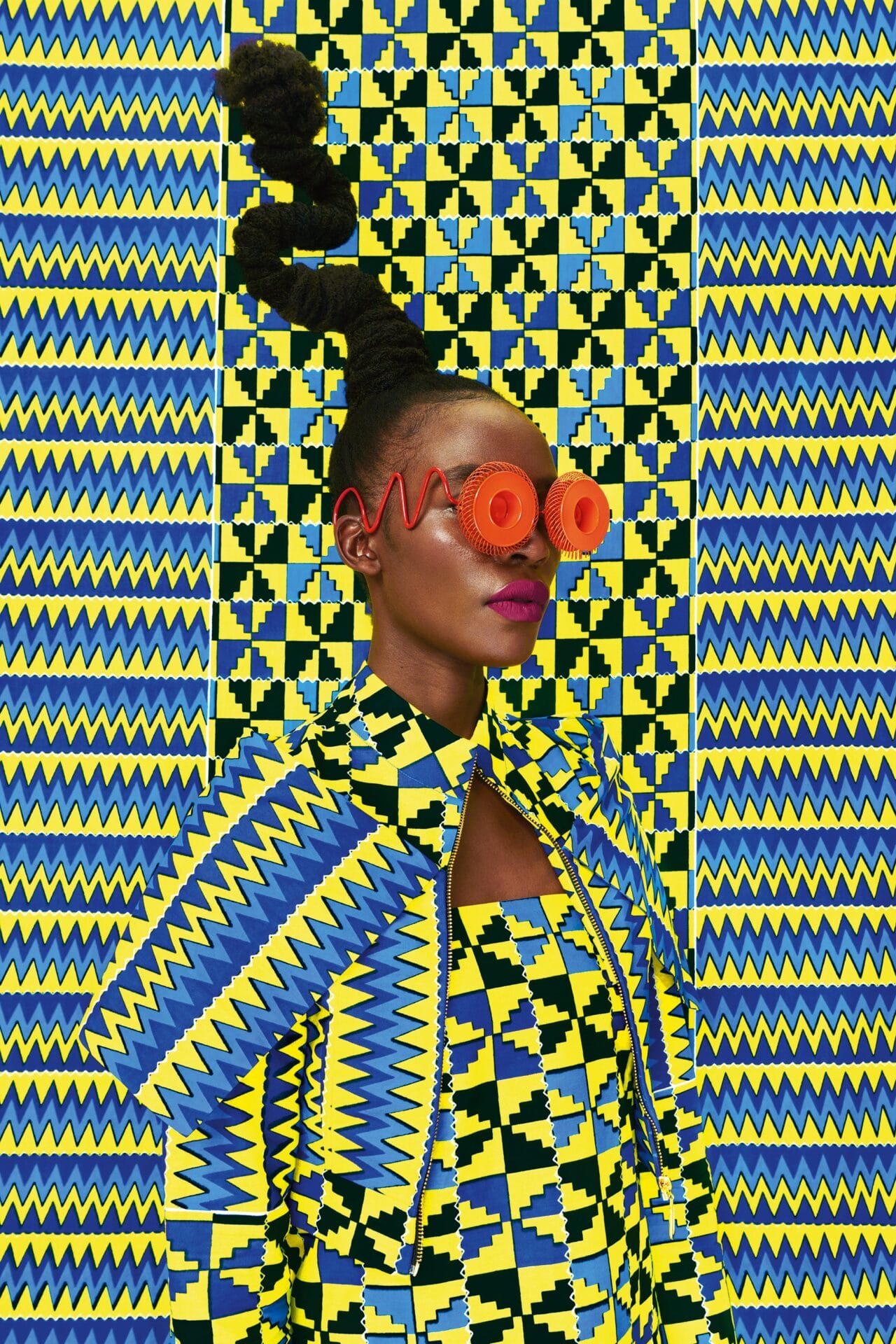 a portrait of the artist against a patterned yellow and blue backdrop. she's wearing bright orange eyewear
