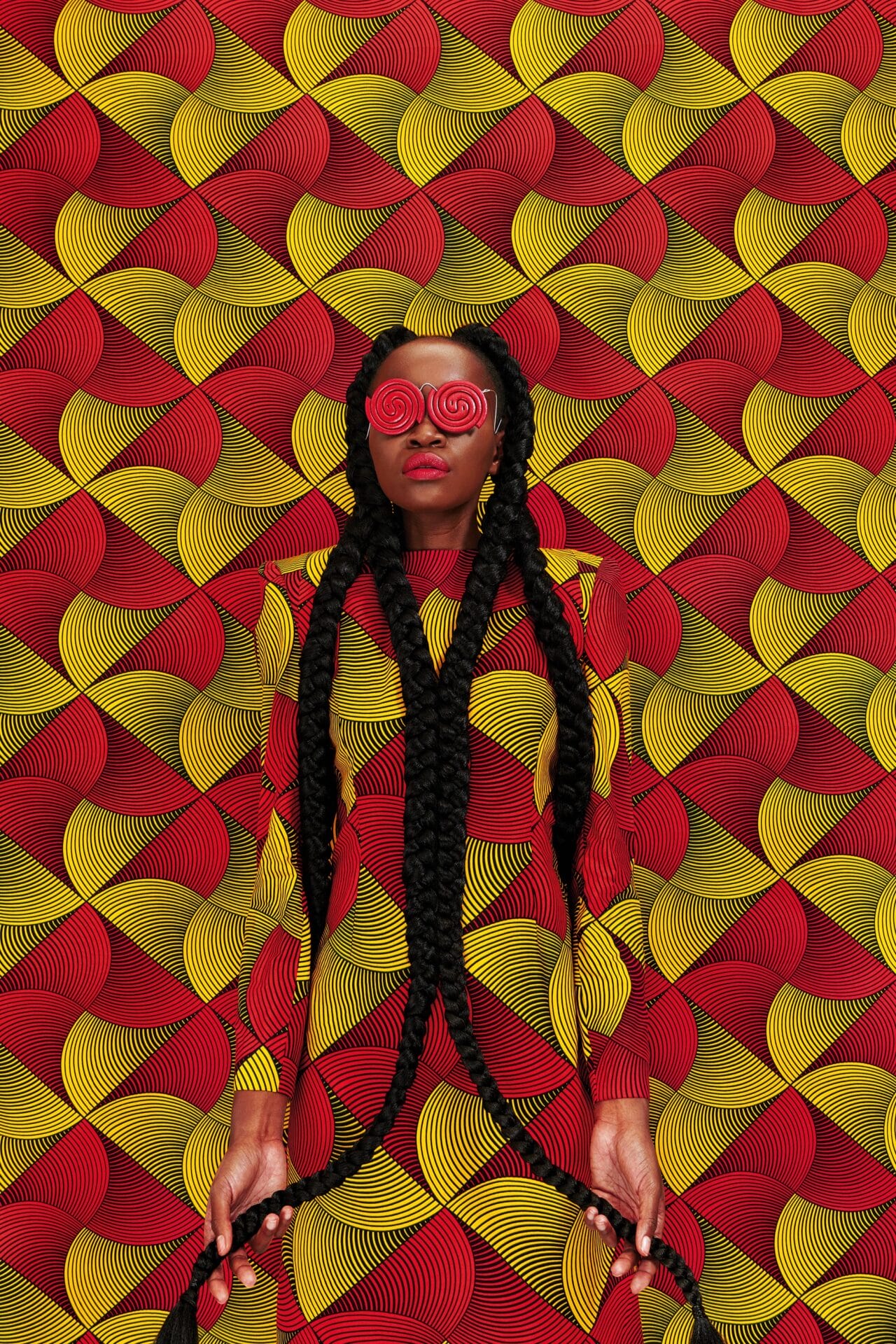 a portrait of the artist against a patterned red and yellow backdrop. she's wearing bright red eyewear