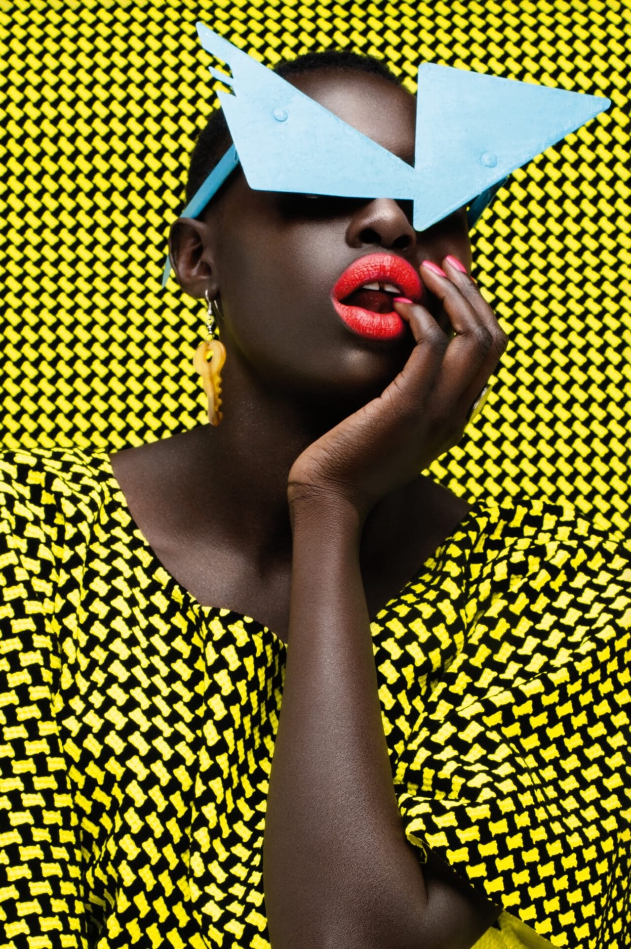 a portrait of the artist against a patterned black and yellow backdrop. she's wearing bright blue eyewear
