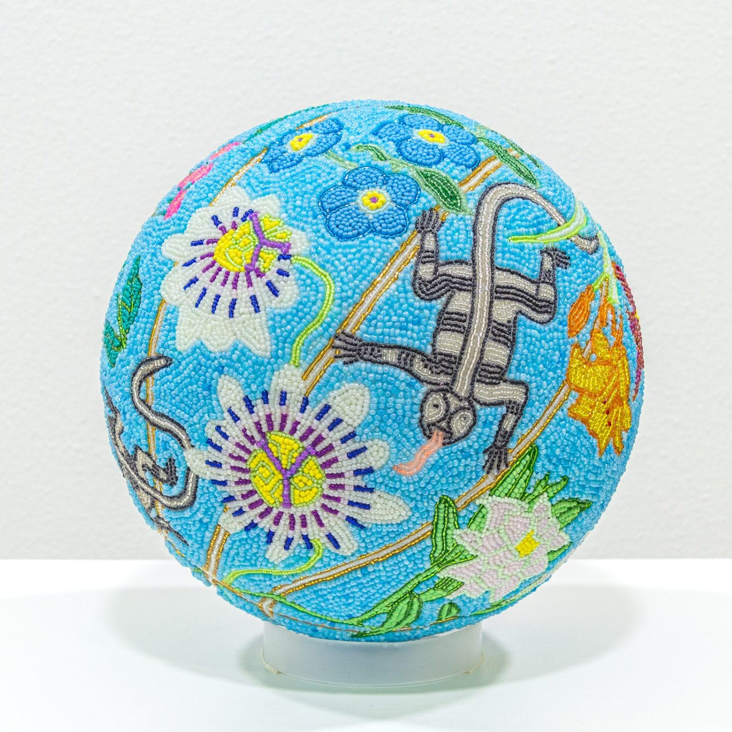 a glass bead-coated spherical sculpture made from a basketball depicting a lizard and numerous kinds of flowers on a blue background