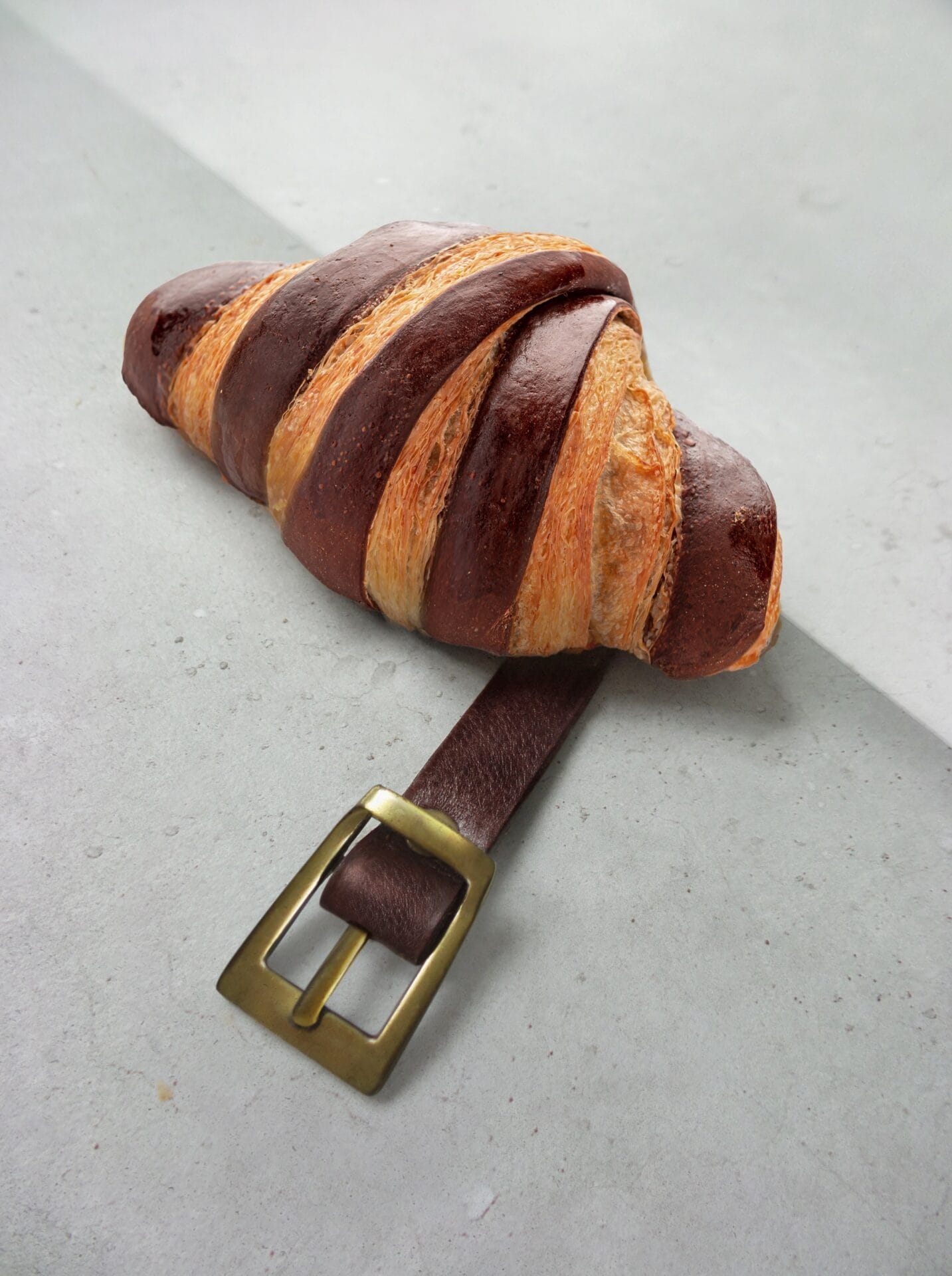 a croissant with dark brown stripes with a belt buckle extending from it as if the belt has been wrapped around the pastry