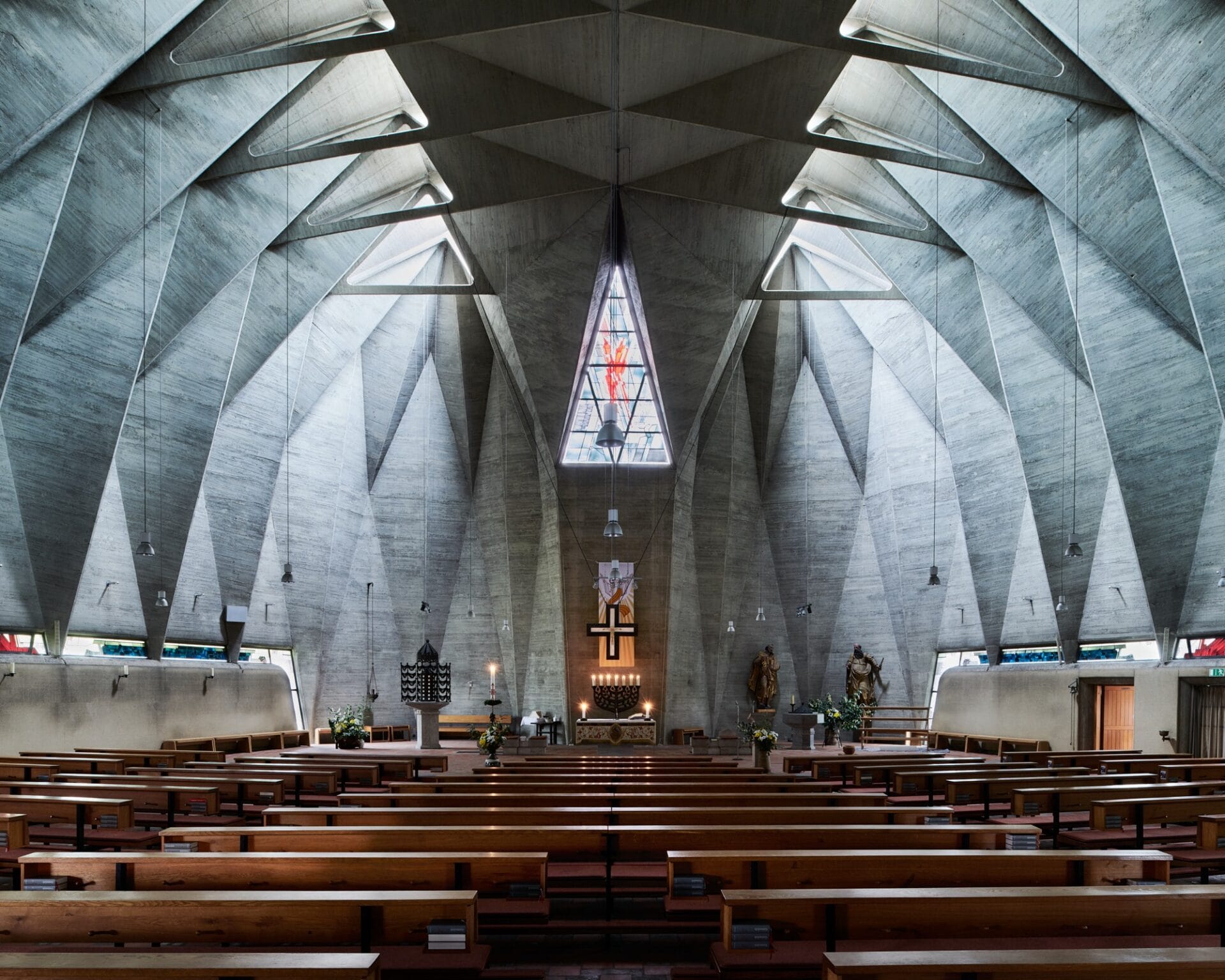 an interior overview of a large brutalist church with angular concrete walls, a clerestory, and wooden pews
