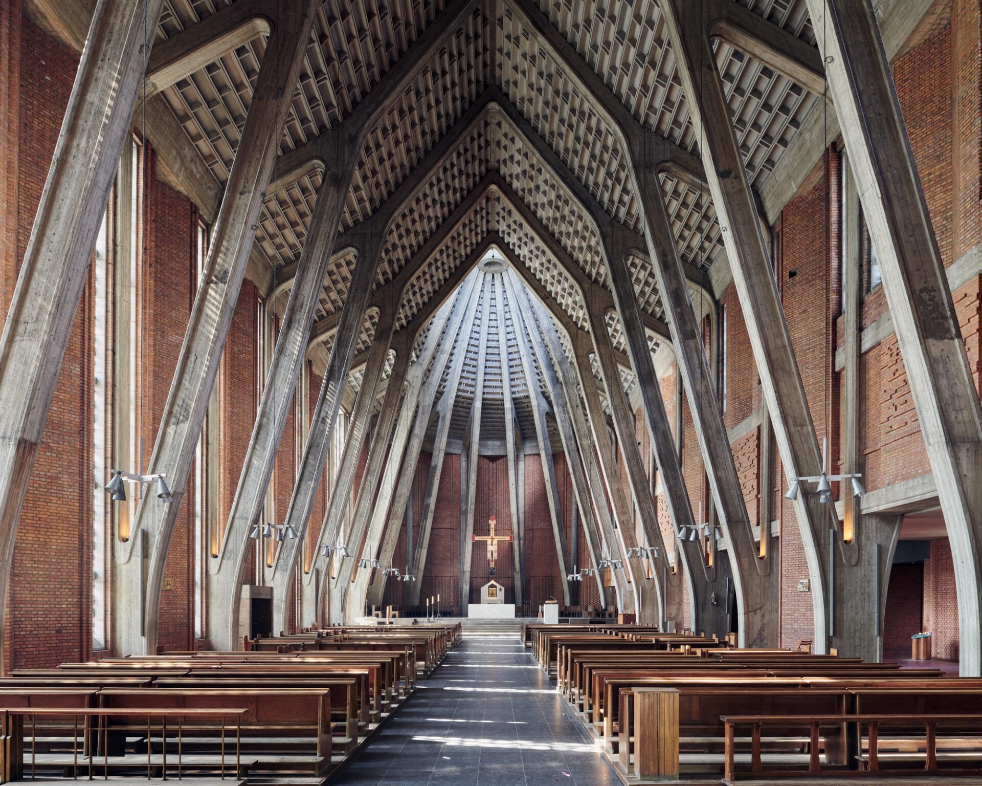 an interior overview of a large brutalist church with brick walls and large, angular concrete and steel beams holding up a pitched ceiling