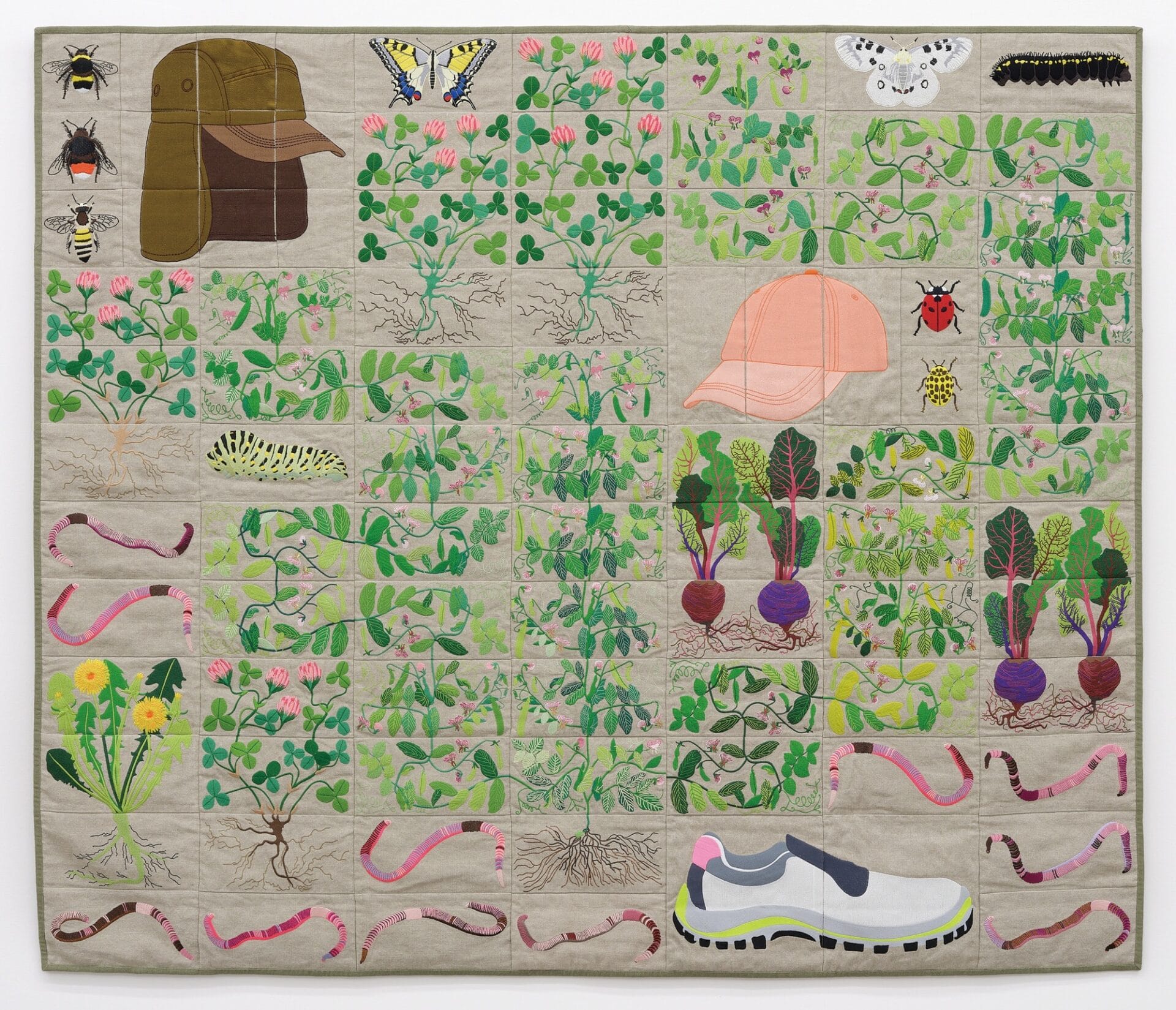 a quilt on a neutral background with plants, worms, insects, two hats, and a shoe