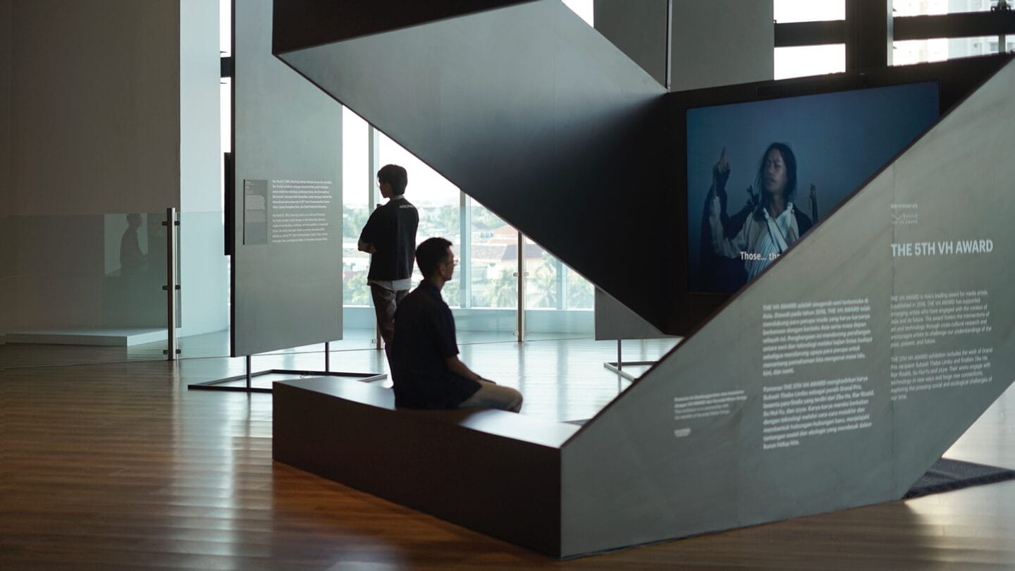 Two people on a platform watching a video (showing a woman) apart of an installation. 