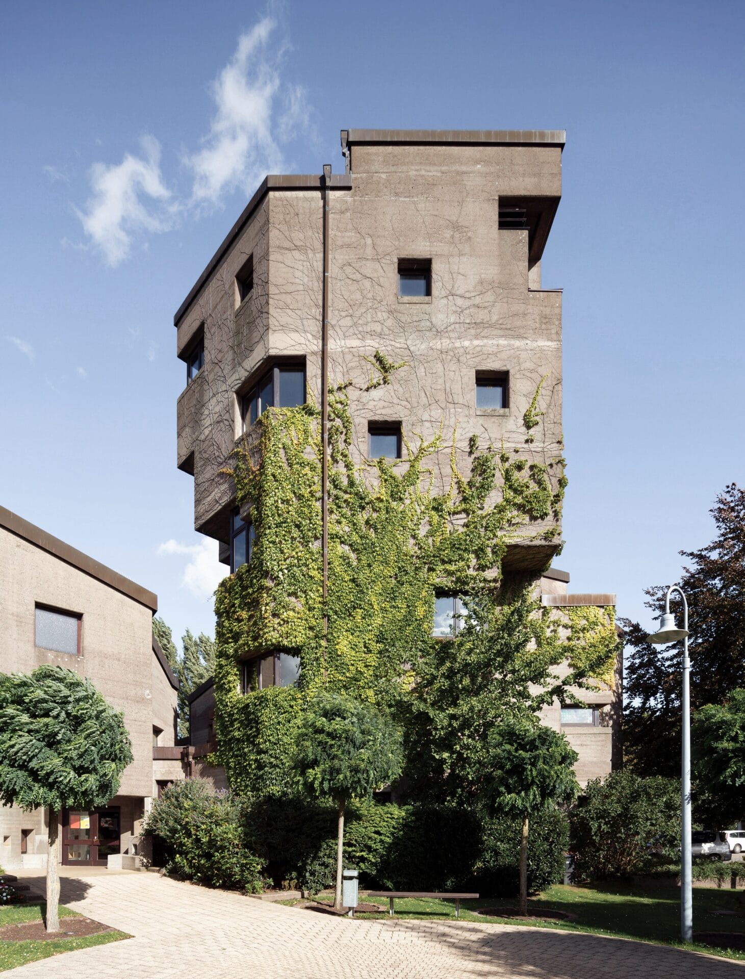 the exterior of a brutalist tower with vines creeping up the wall and trees around the base
