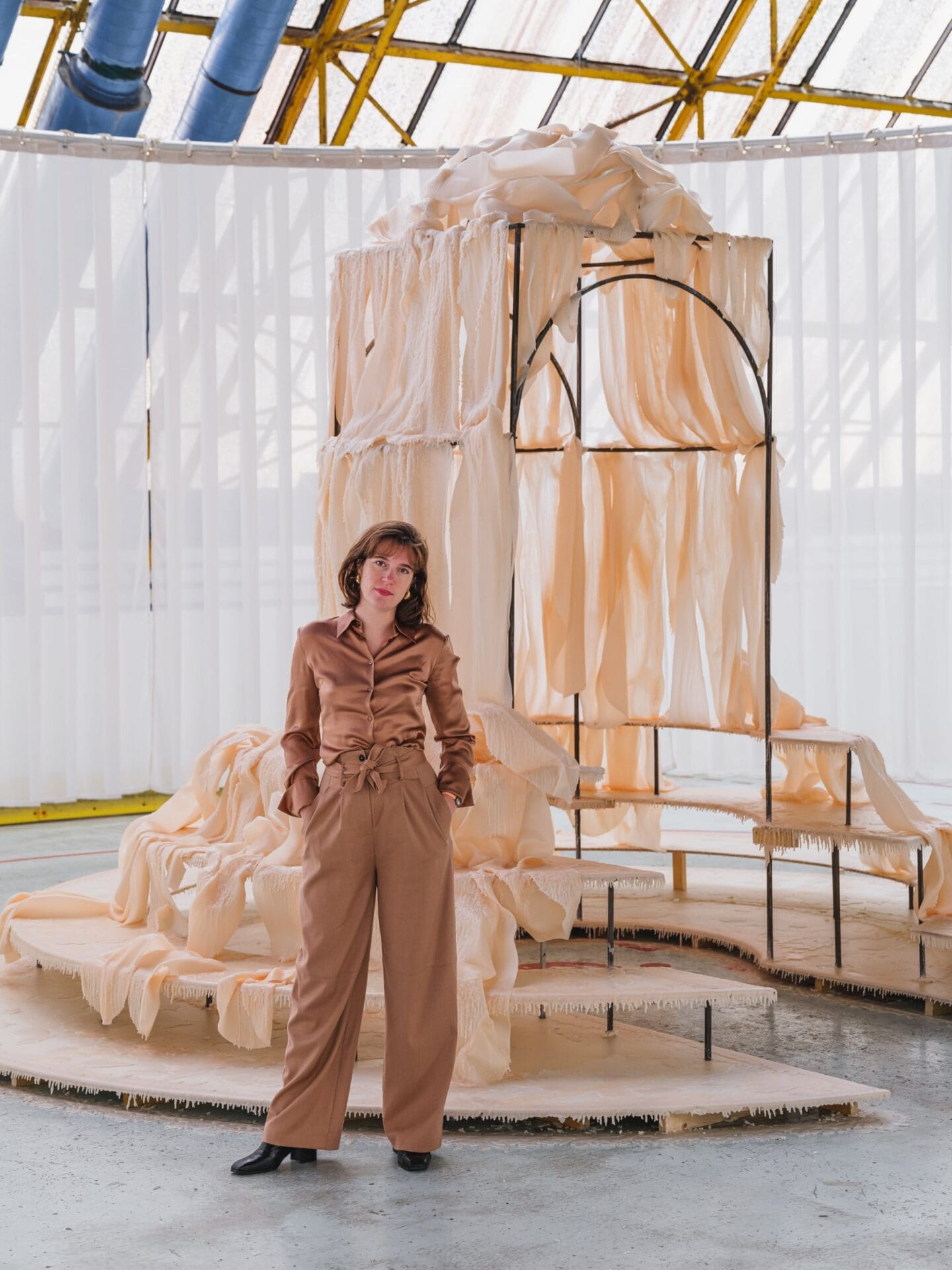 a white woman with red hair in a beige button up and pants stands in front of a tall architectural armature draped with wax sheets
