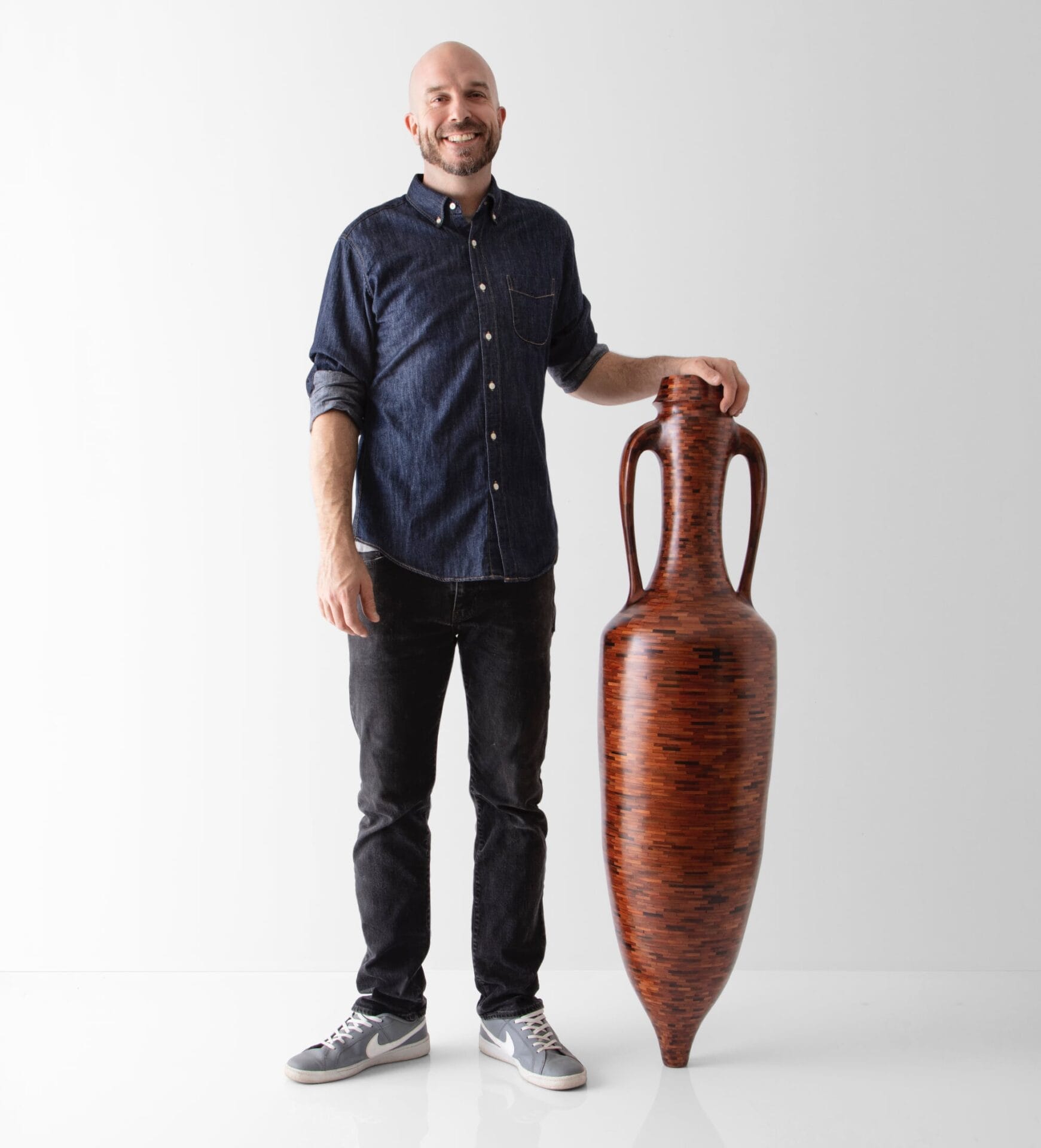 a bald white man in a jean shirt, jeans, and sneakers stands next to a tall wooden amphora