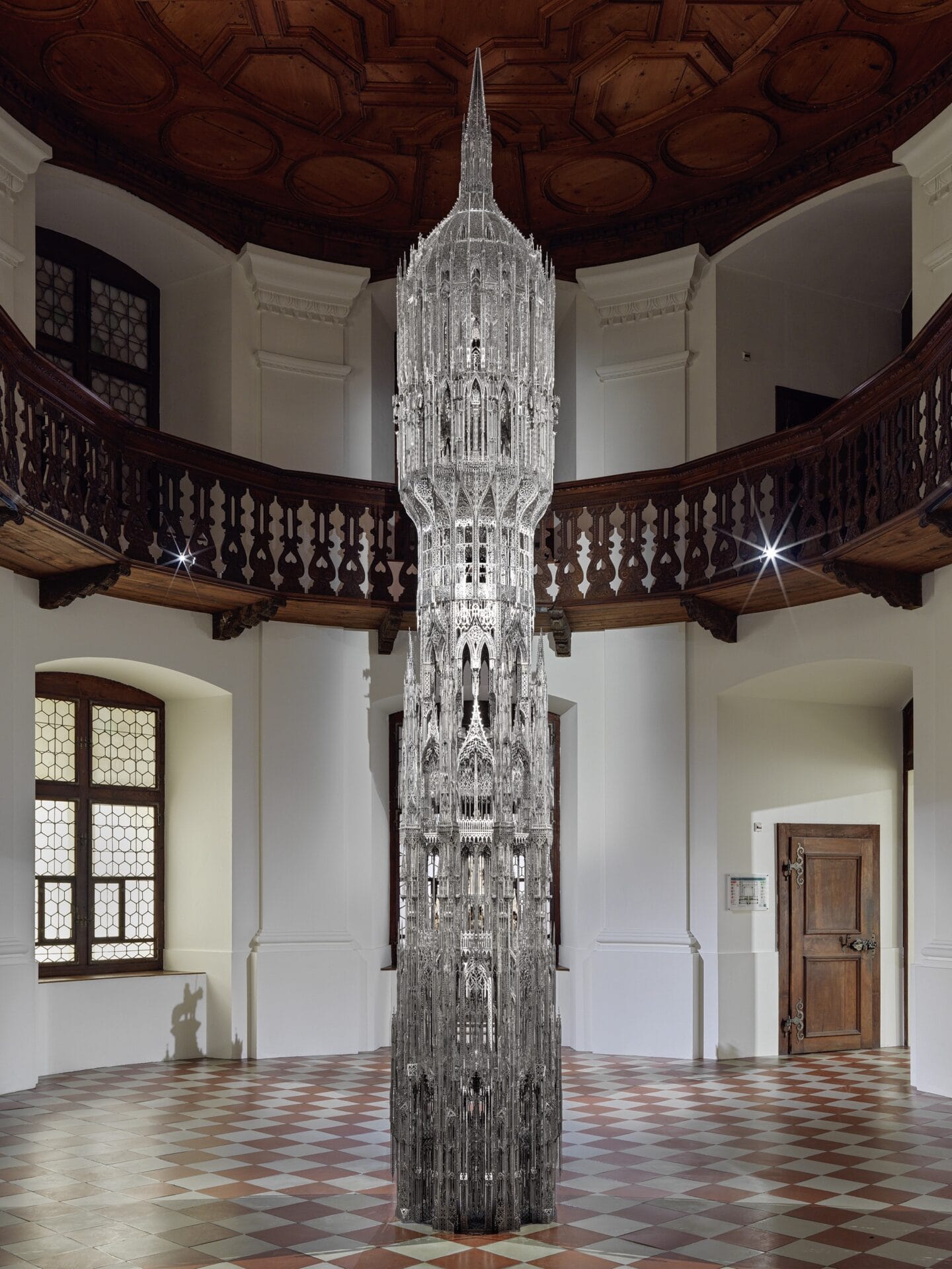 a tall metallica sculpture in the middle of a historic gallery with a wooden mezzanine in the background