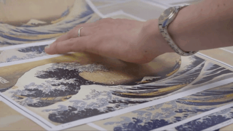 a gif from a short documentary about Hokusai's "The Great Wave" print, showing hands moving scans of the print around on a table