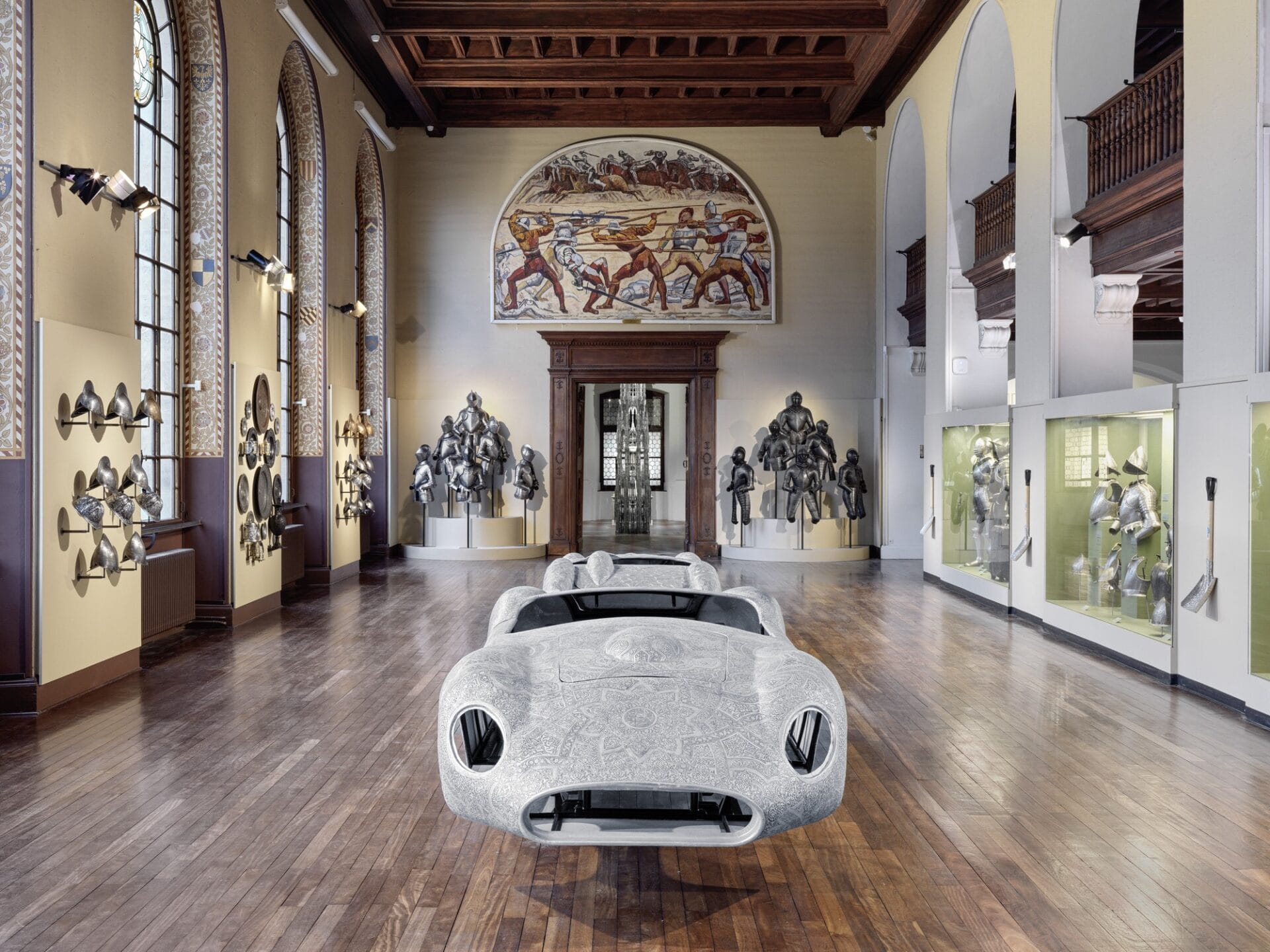 an installation view in a museum showing a sculpture of a shell of a sports car surrounded by walls of armor