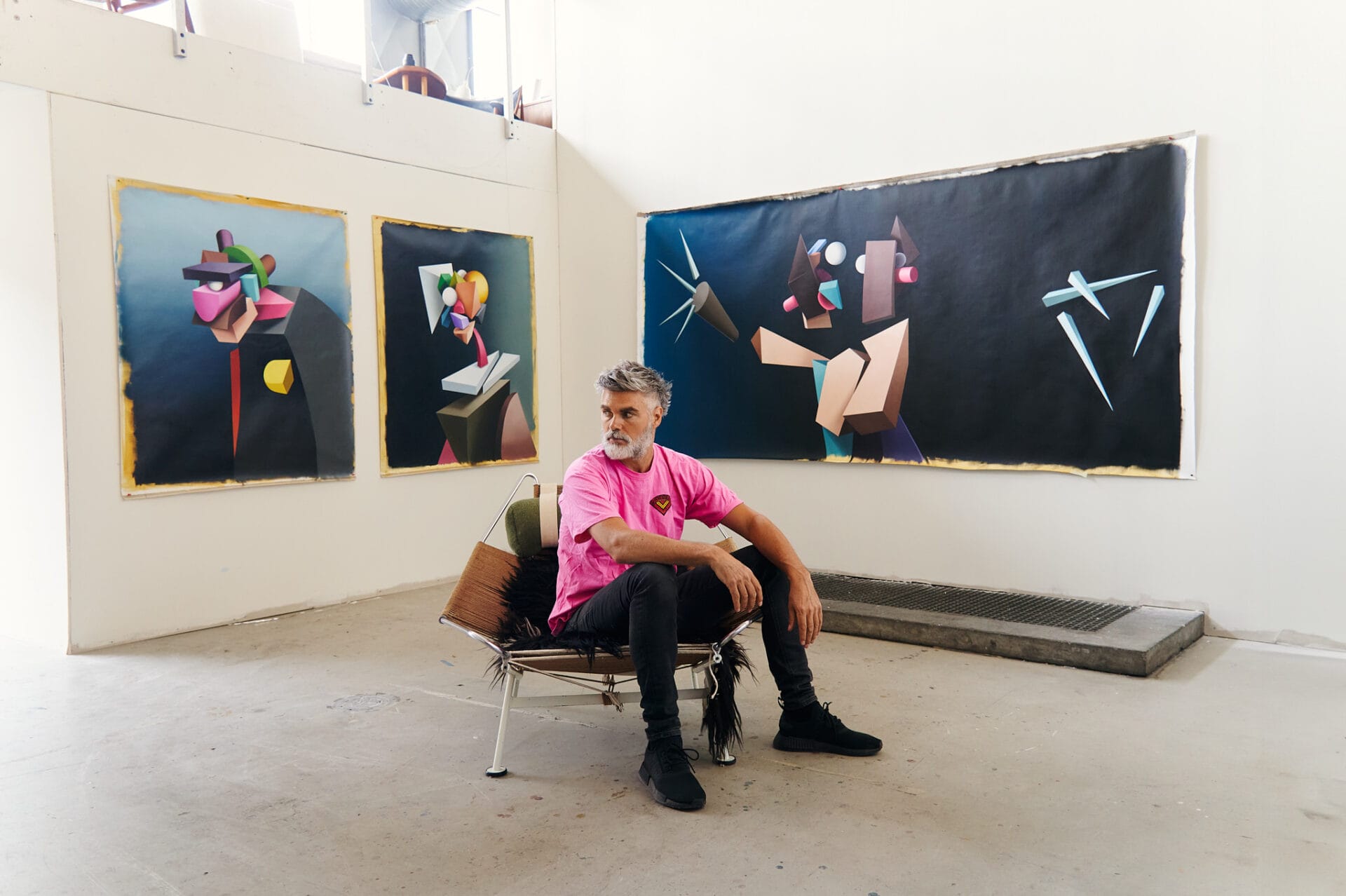the artist wears a pink shirt and black pants and sits on a fur-covered chair in a studio with three portraits lining the walls