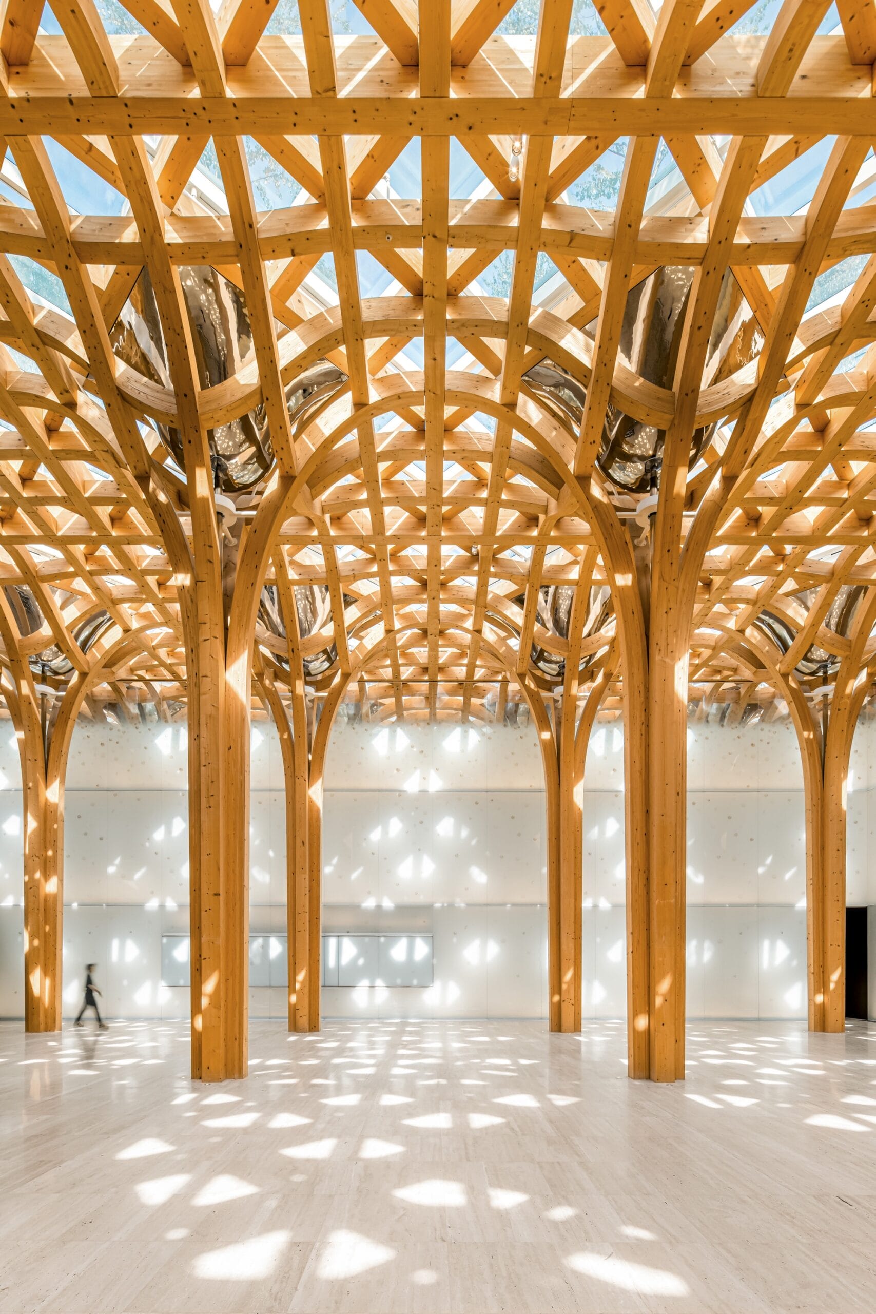 a vertical image of the interior of a sunlit architectural space with white floors and walls and intricate wooden latticework on the ceiling that blends into a series of support columns