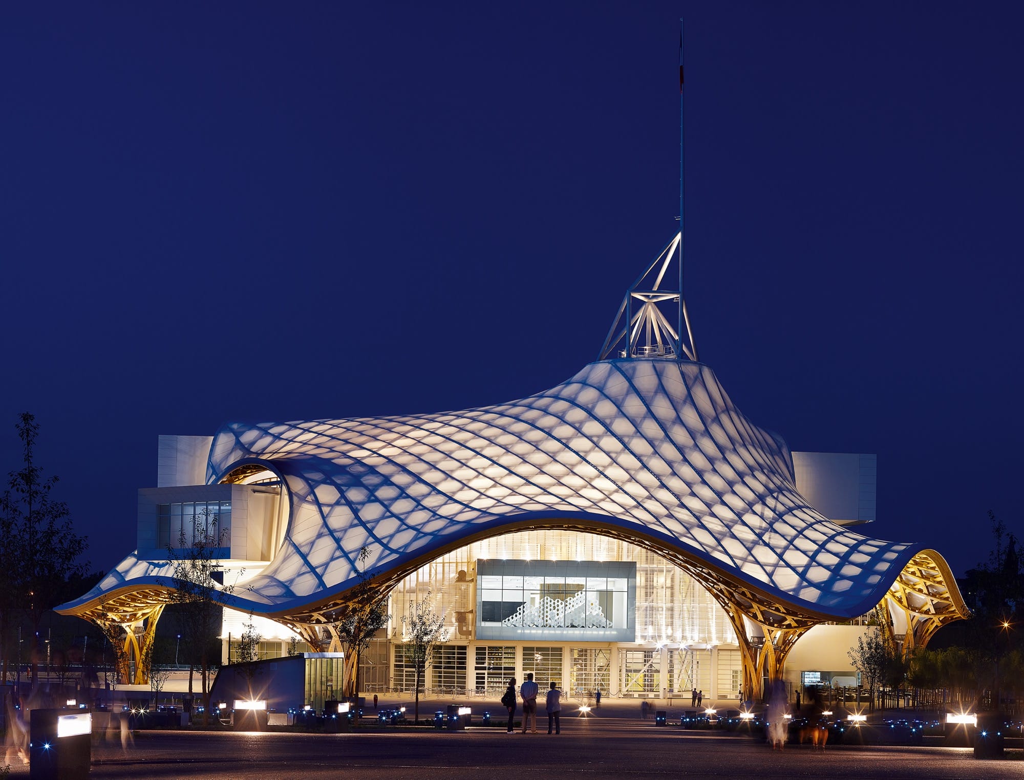 a striking, undulating building viewed from outside at night, with light coming through latticework in the ceiling