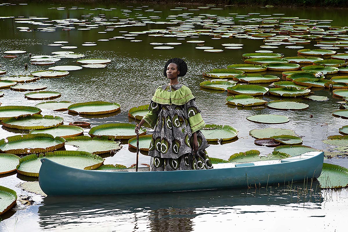 a portrait photograph of a Black woman wearing a Dutch wax fabric dress, standing in a blue canoe among numerous large lily pads