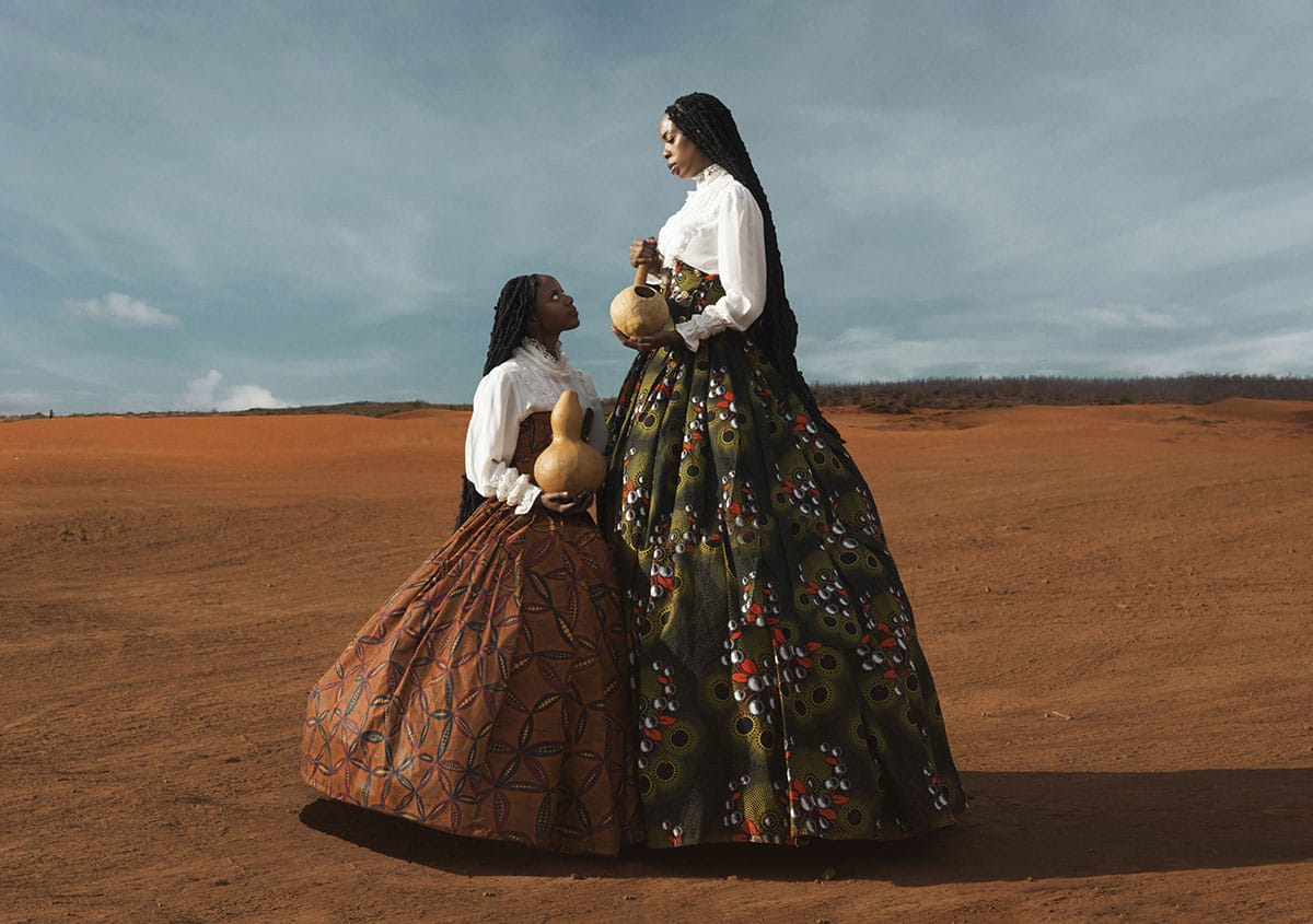 a photograph of two woman standing facing one another in a desert, both wearing dresses and holding gourds, one much taller than the other