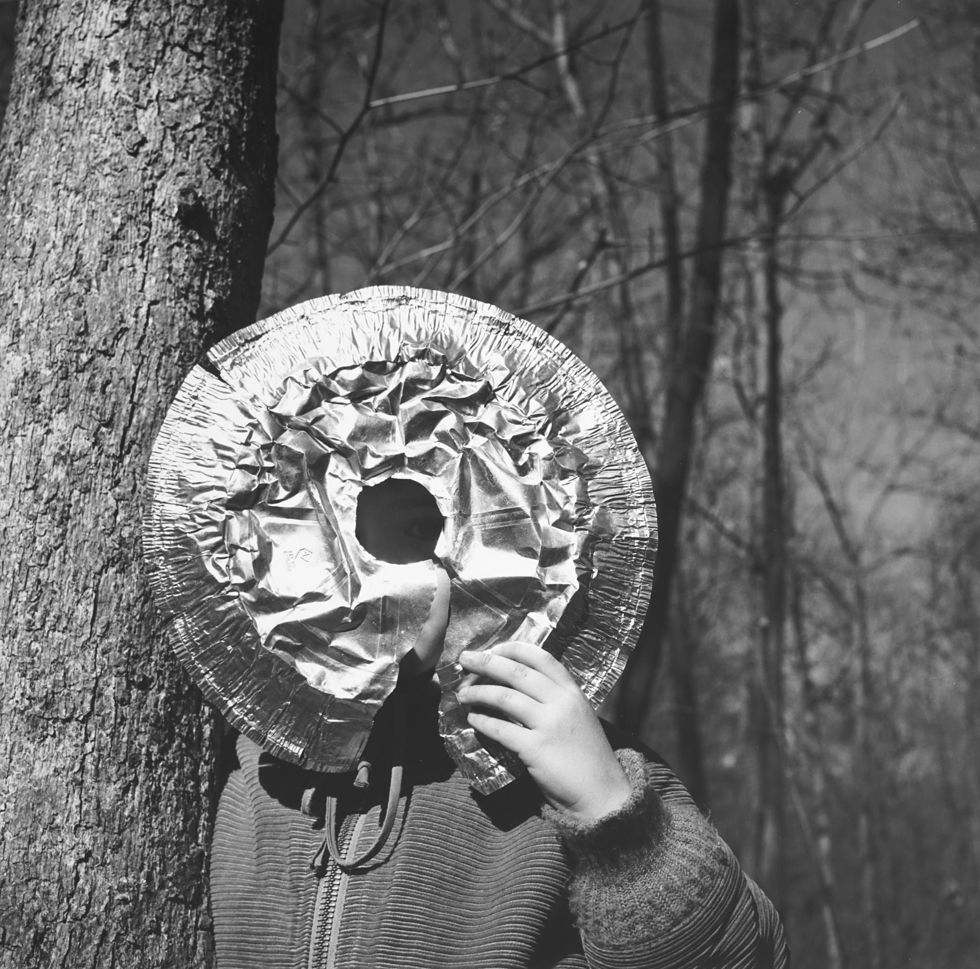 a black and white photo of a child holding an aluminum pie tin up to their face and looking through a hole in the middle. they're in a forest