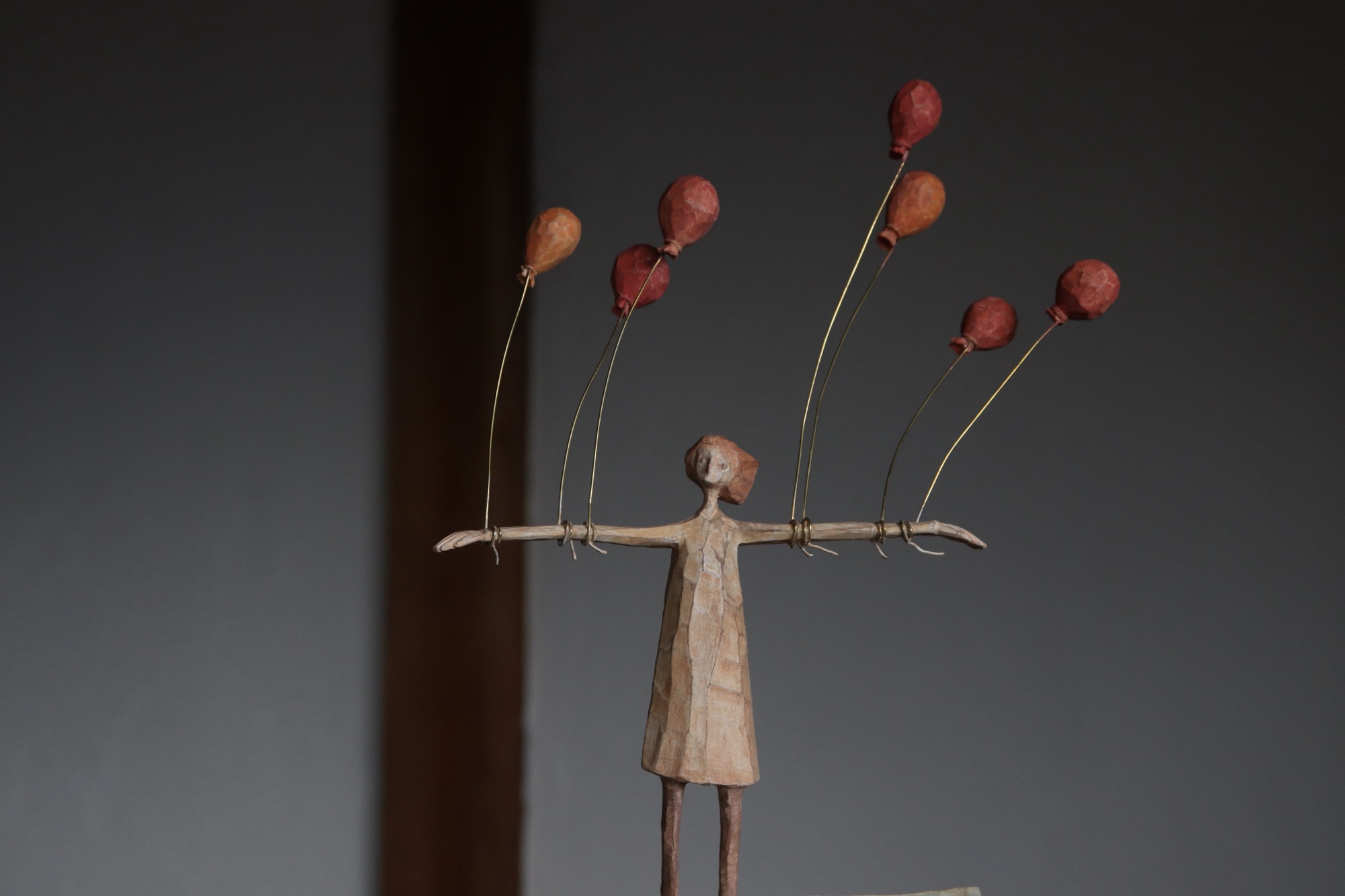 a wooden sculpture of a slender figure with arms outstretched with balloons tied to them