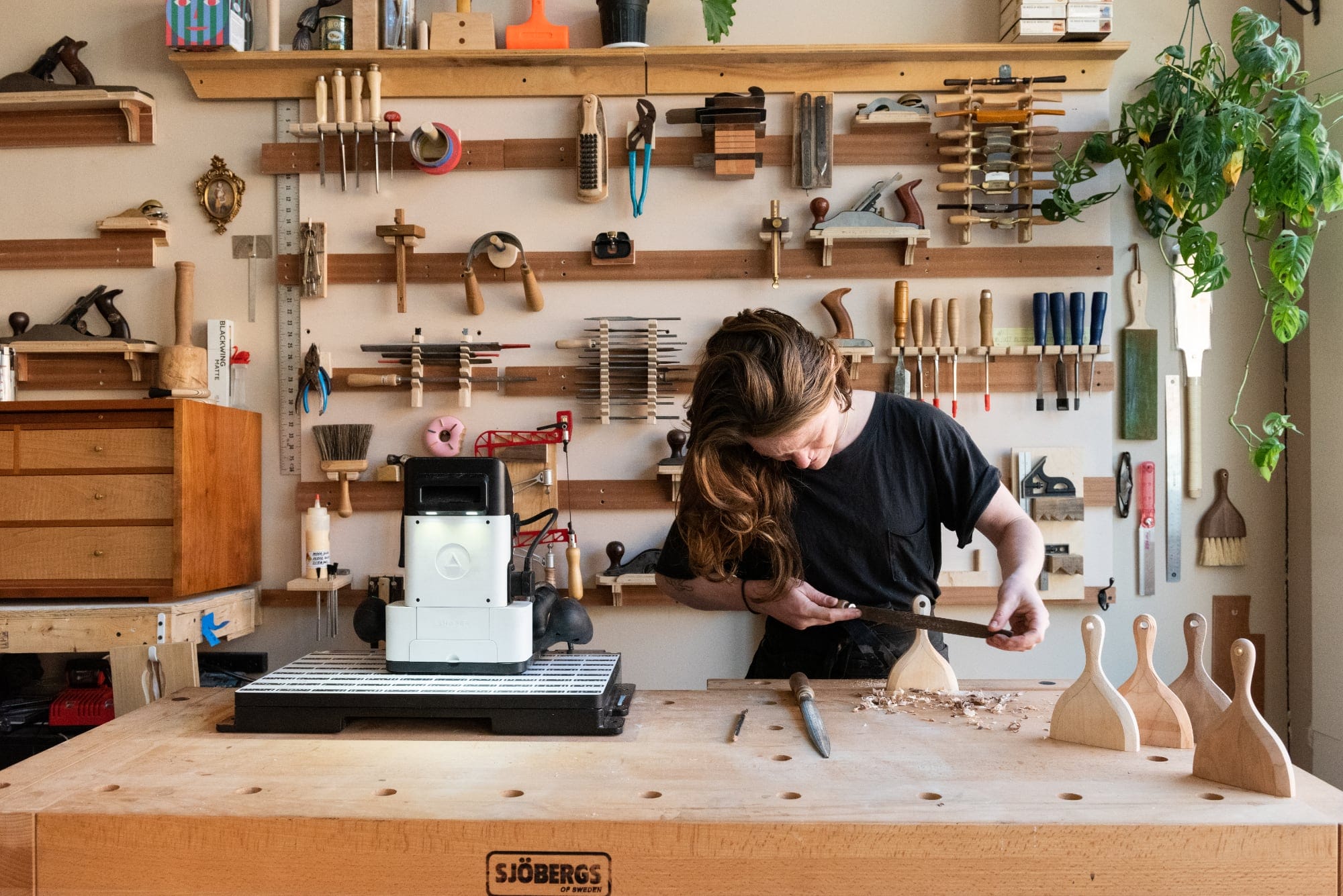 the artist carving a wooden handle at a table in a woodshop with tools on the wall behind her