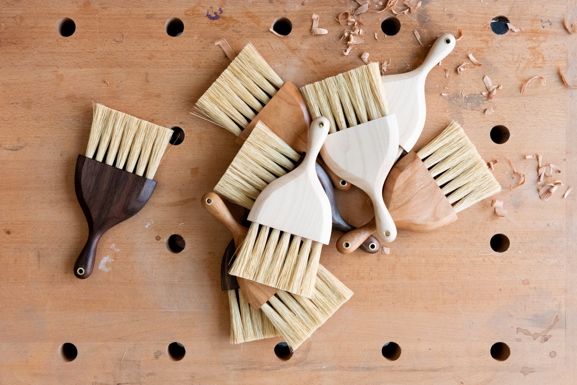 a pile of small hand-held brushes made with a variety of woods on a wooden surface with holes and wood shavings