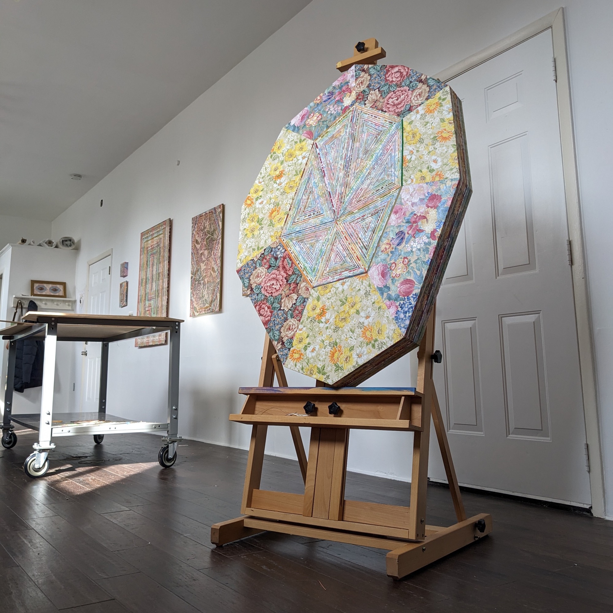 an artwork on a shaped panel sits on an easel in a studio, with other artworks on the wall in the background and a work table nearby
