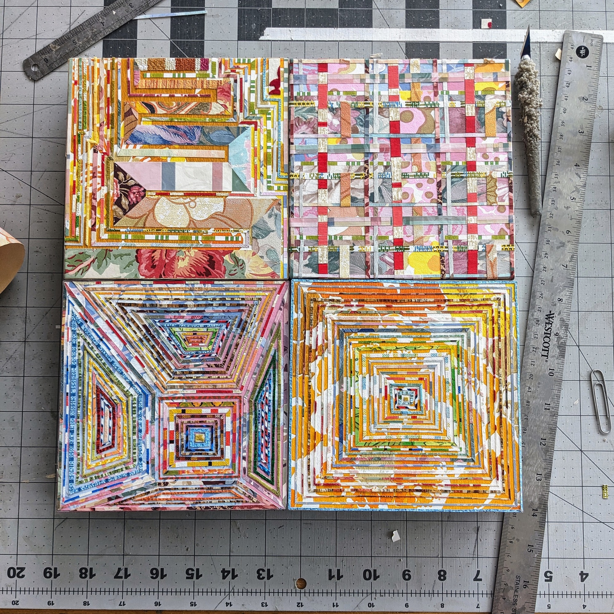 four small, square artworks made from strips of vintage wallpaper, grouped together on a work table with a ruler and other tools nearby