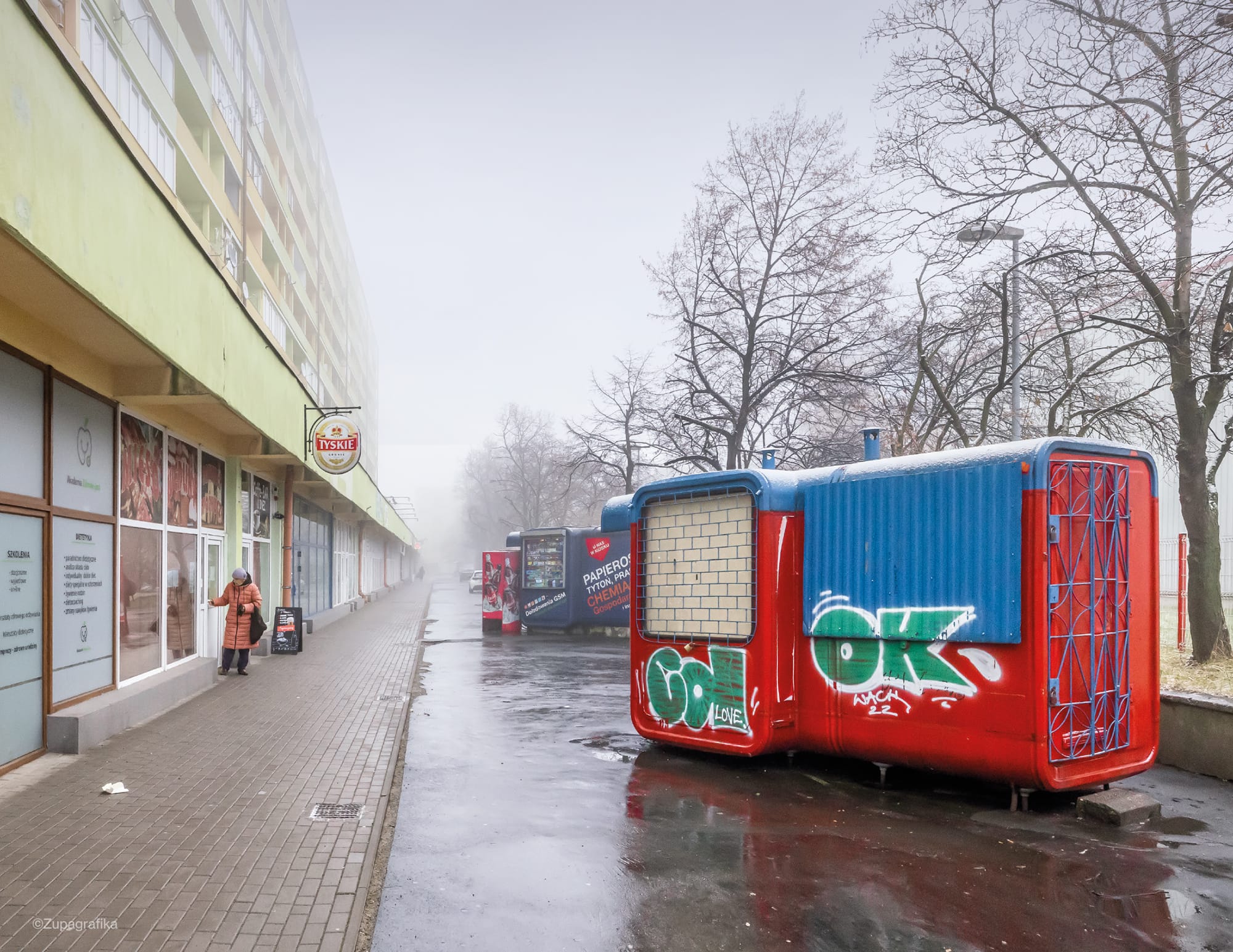 a photograph of a series of modernist kiosks outside of a shopping center on a foggy day in Poland. the kiosk in the foreground has green graffiti on it that reads "OK"