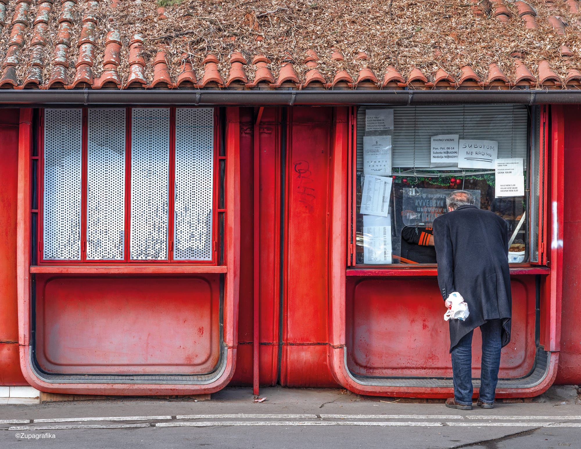 a man peers into the window of a red, modernist, modular kiosk with a Spanish tile roof