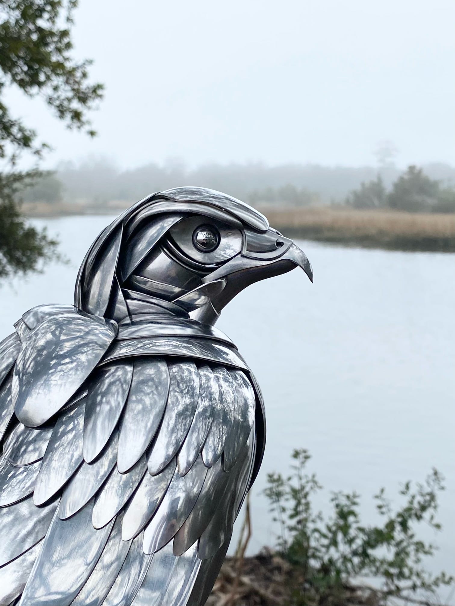 a metal bird made from repurposed silverware perches in the trees, a body of water rests in the background