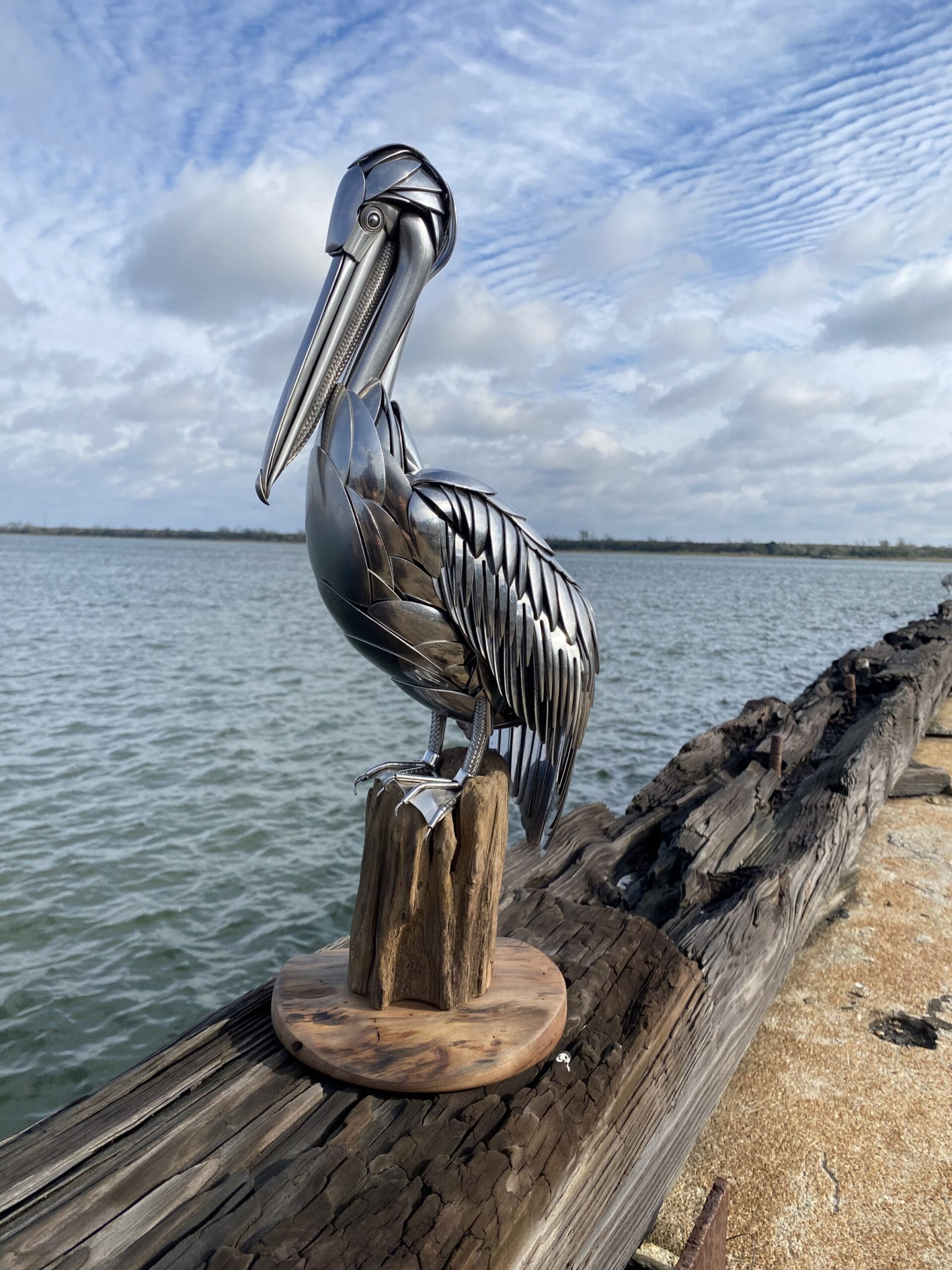 a metal bird made from repurposed silverware perches upon a wooden post next to a lake