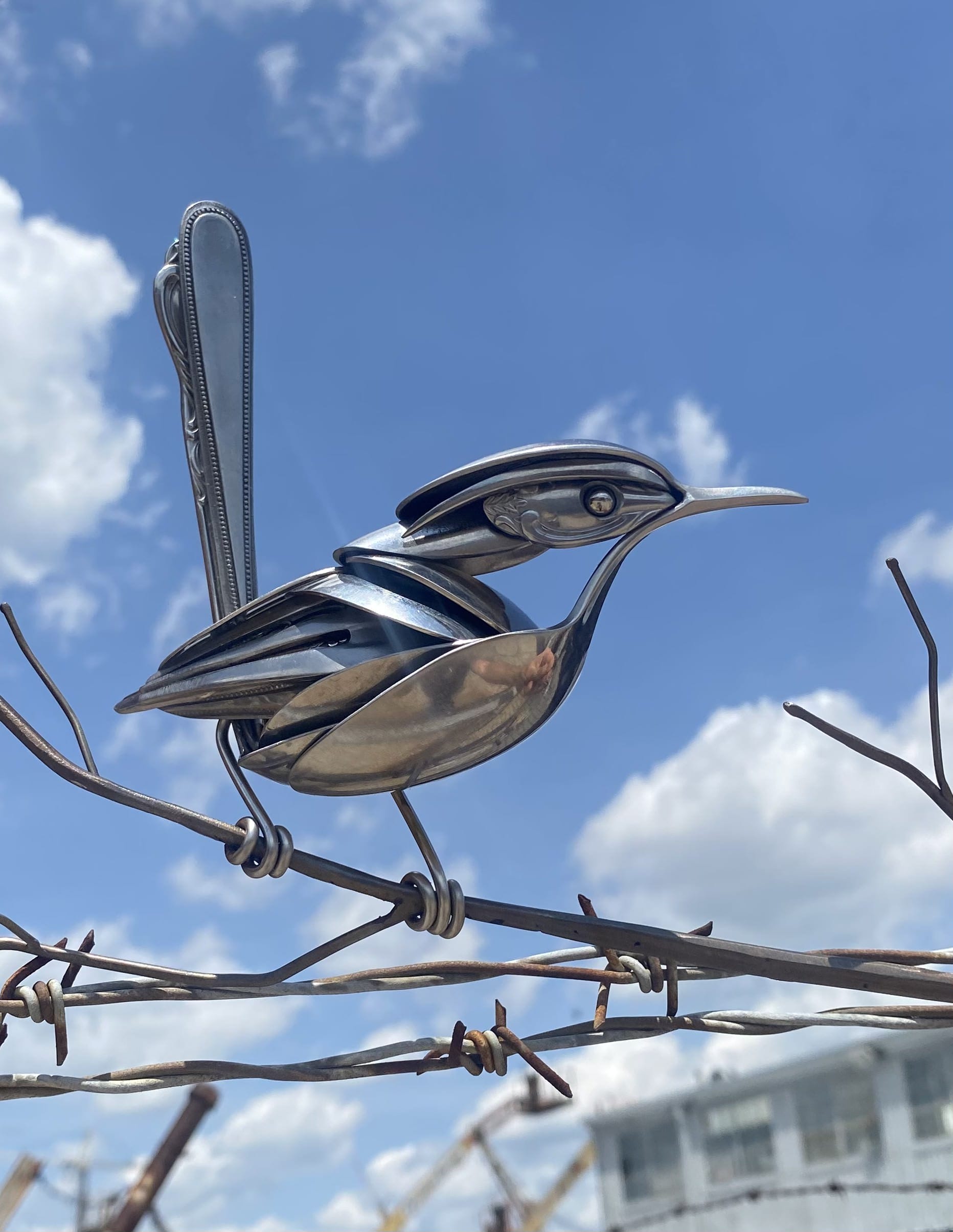 a metal bird made from repurposed silverware perches upon a metal branch resembling barbed wire