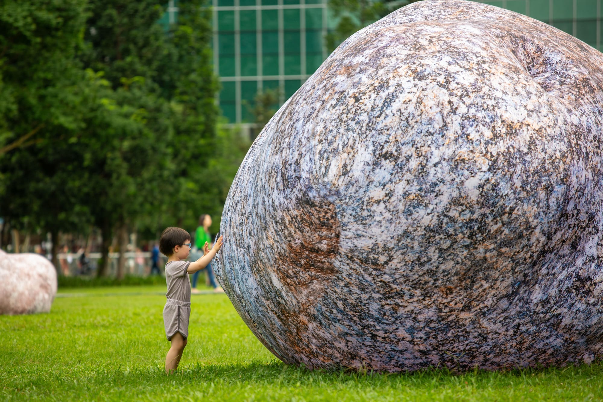 a small child reaches outward at a large boulder-looking form in a park