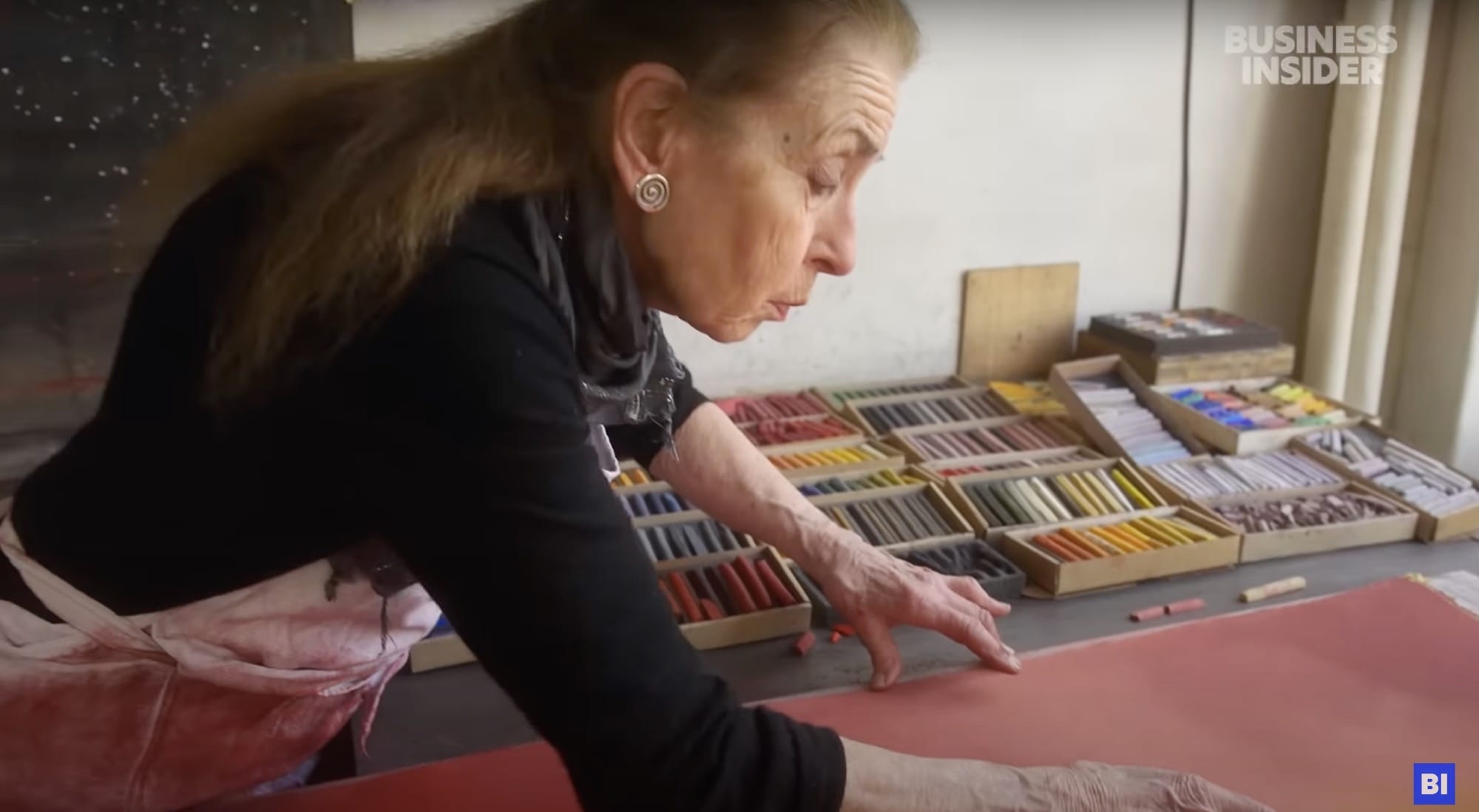 a still from a short documentary about a pastel maker, showing an artist working on a drawing with boxes of pastels in the background