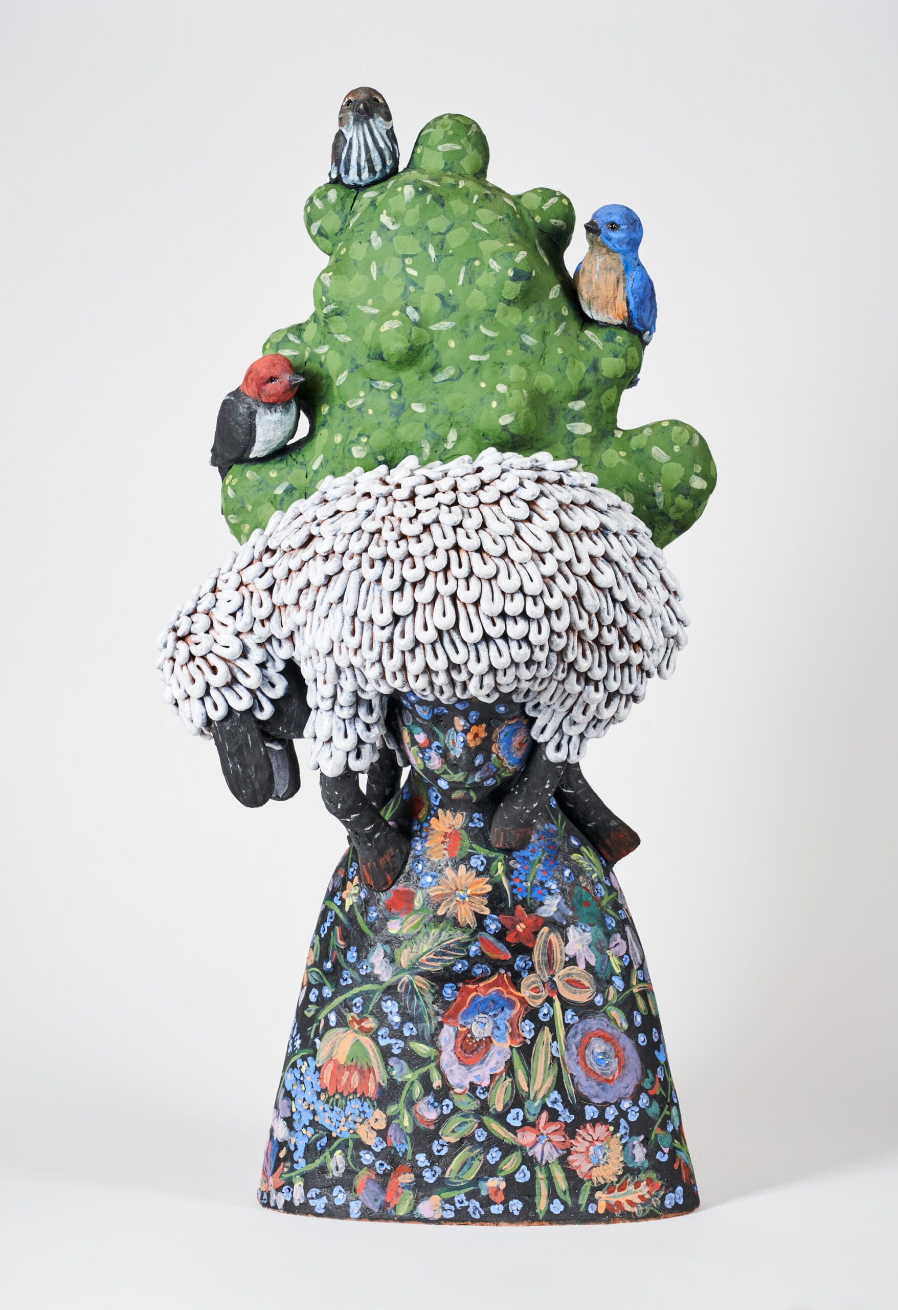 a ceramic sculpture of a figure covered in floral motifs with a lamb on its head. a tree with three birds nesting in it ascends upward.