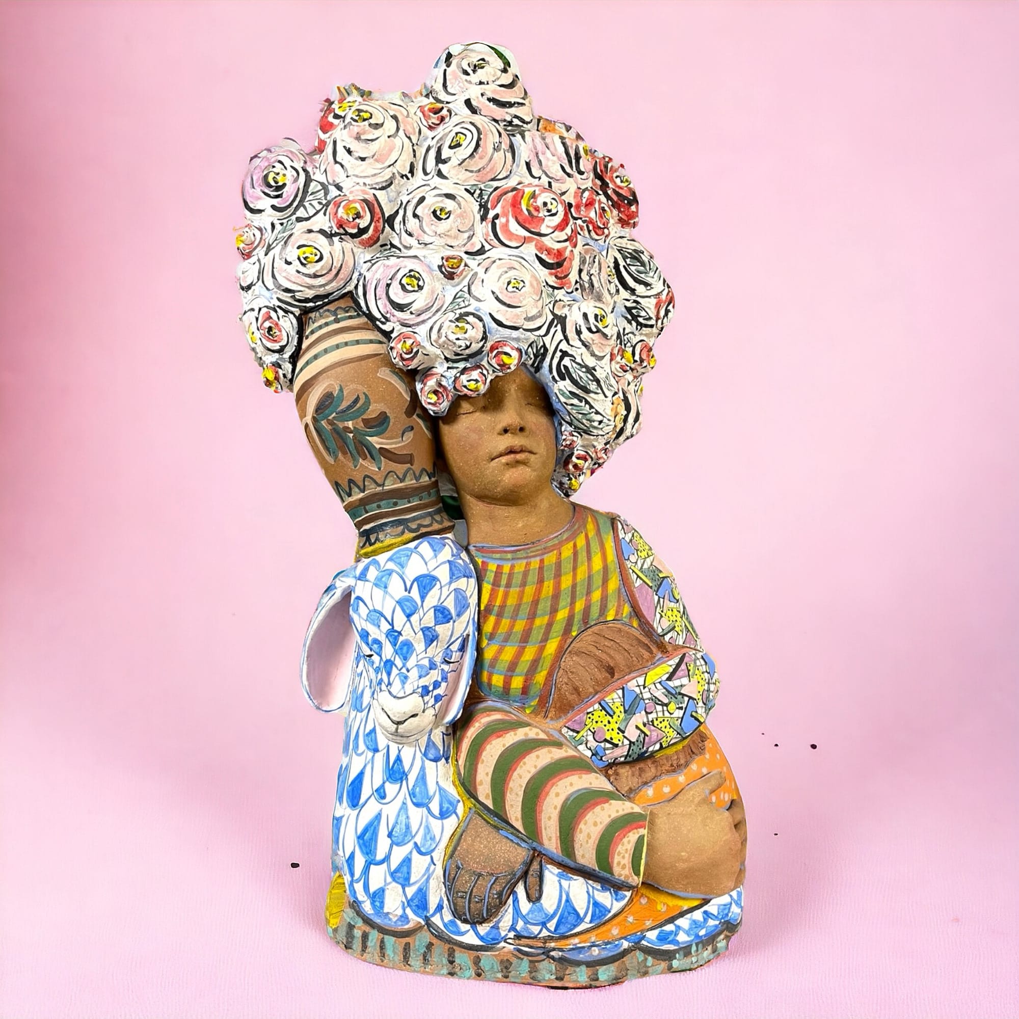 a ceramic sculpture of a figure with its eyes closed, embracing both a lamb and another individual. a throng of flowers covers the top of the composition.