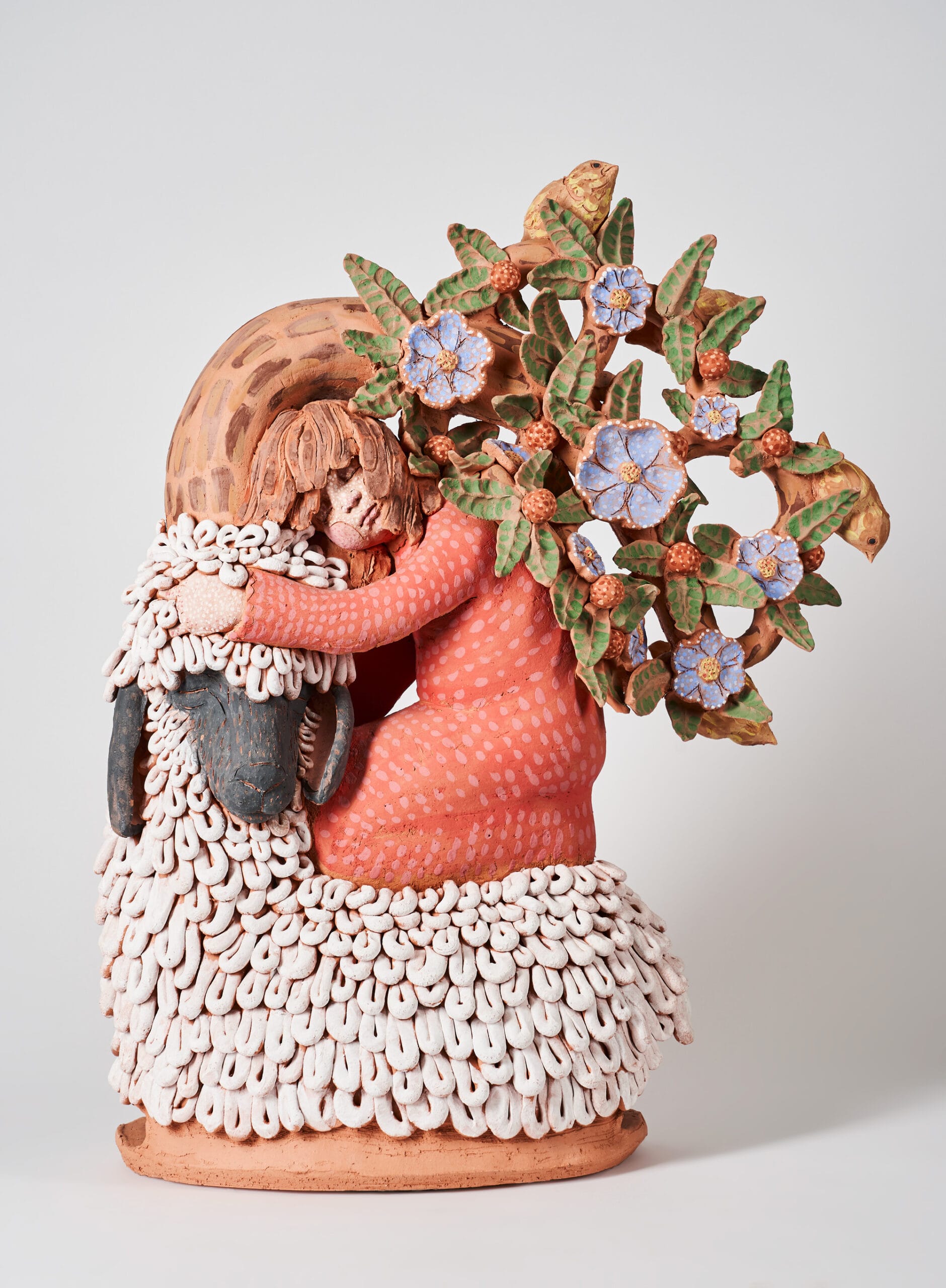 a ceramic sculpture of a young girl embracing a lamb. a tree bends behind them, almost as if it is embracing the small girl as well.