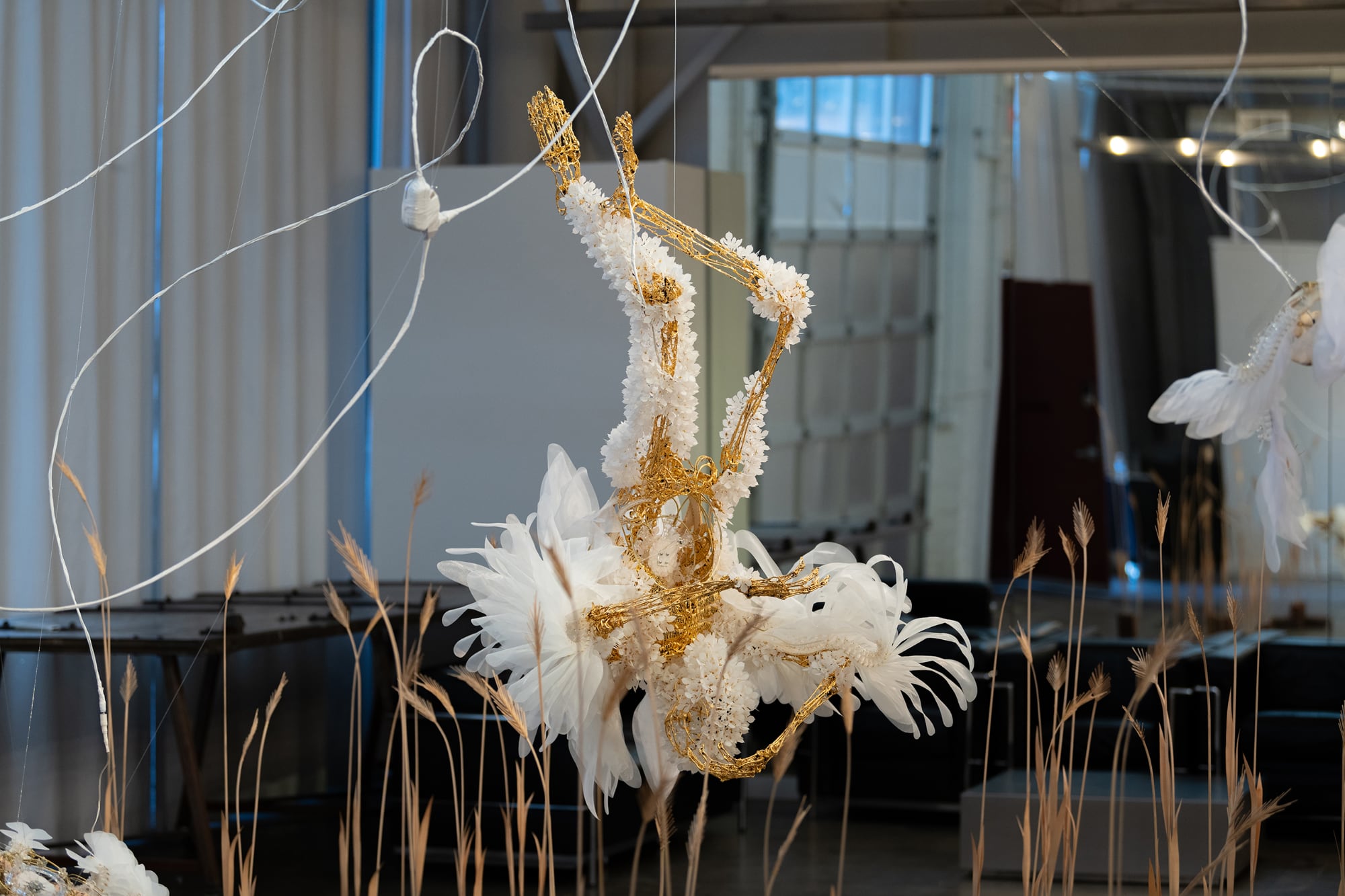 a figurative sculpture with a gold skeleton and white fluttery wings hangs upside down in a gallery with wheat on the floor