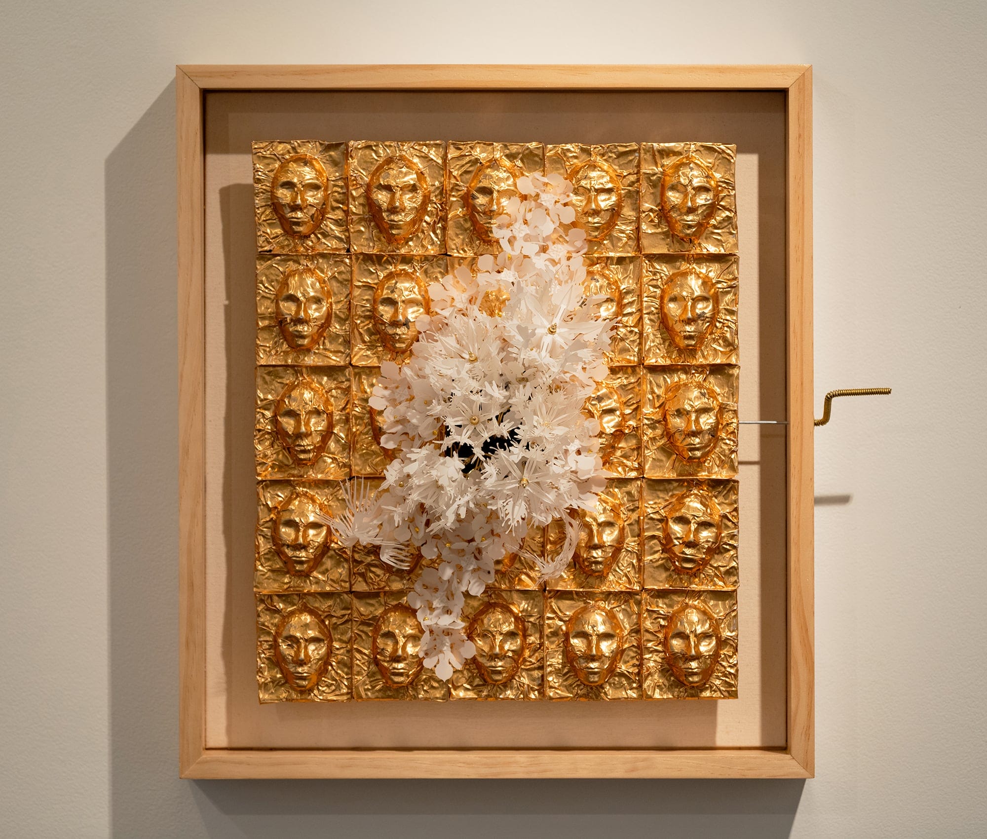 a grid of gold faces with a white fluttering mass on top in a wooden frame with a hand crank to the right