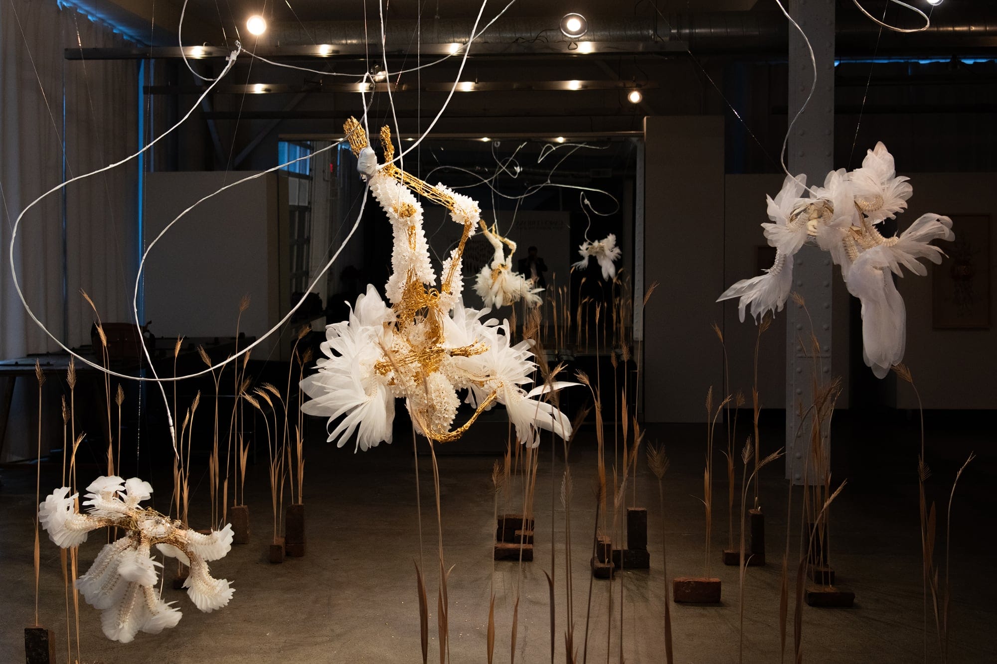 an installation view of three suspended white and gold sculptures with small sprigs of wheat on the floor