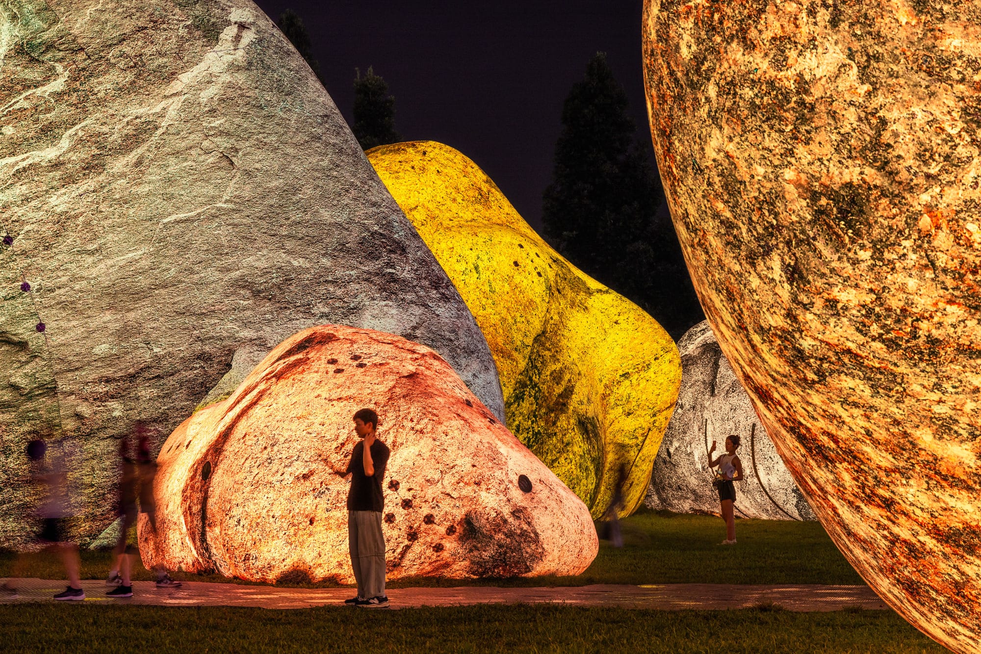 towering rocks rest in a park in glowing hues of peach, tan, and yellow. passerby observe.