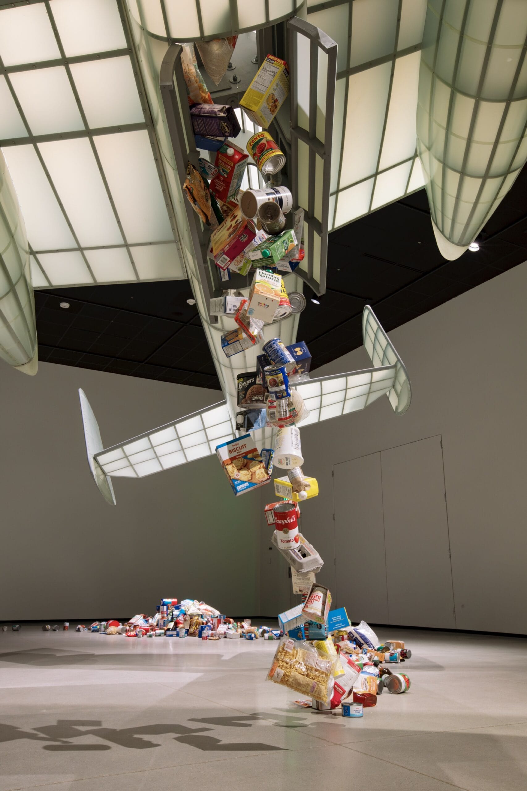 the rear view of a sculpture installation in a large gallery space of a scaled-down B-25 bomber releasing a stream of food onto the ground in its path