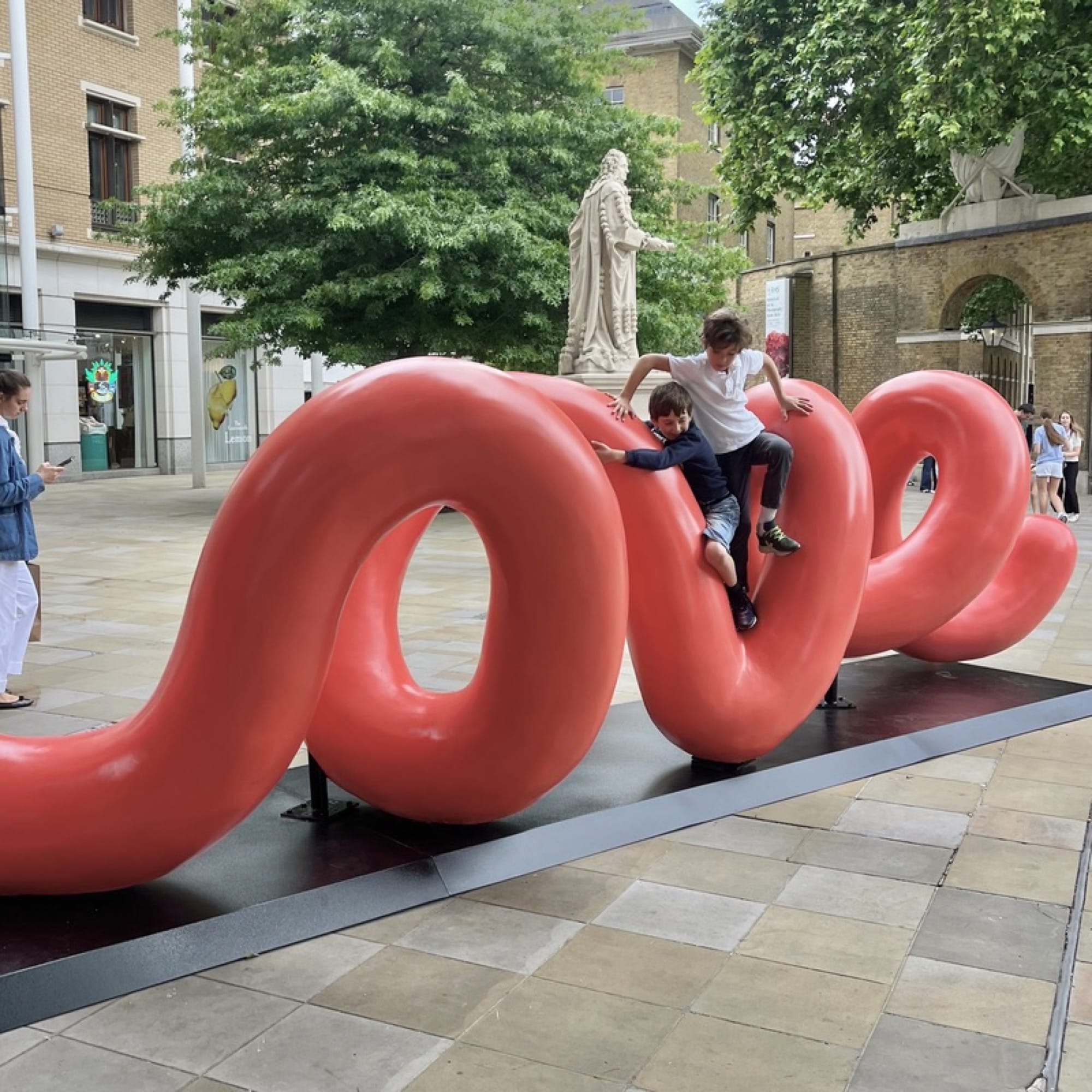 a large-scale, red public sculpture of curving lines that spell the word "love" when viewed from the right perspective, shown in this image with children climbing on top of it in London's Duke of York square