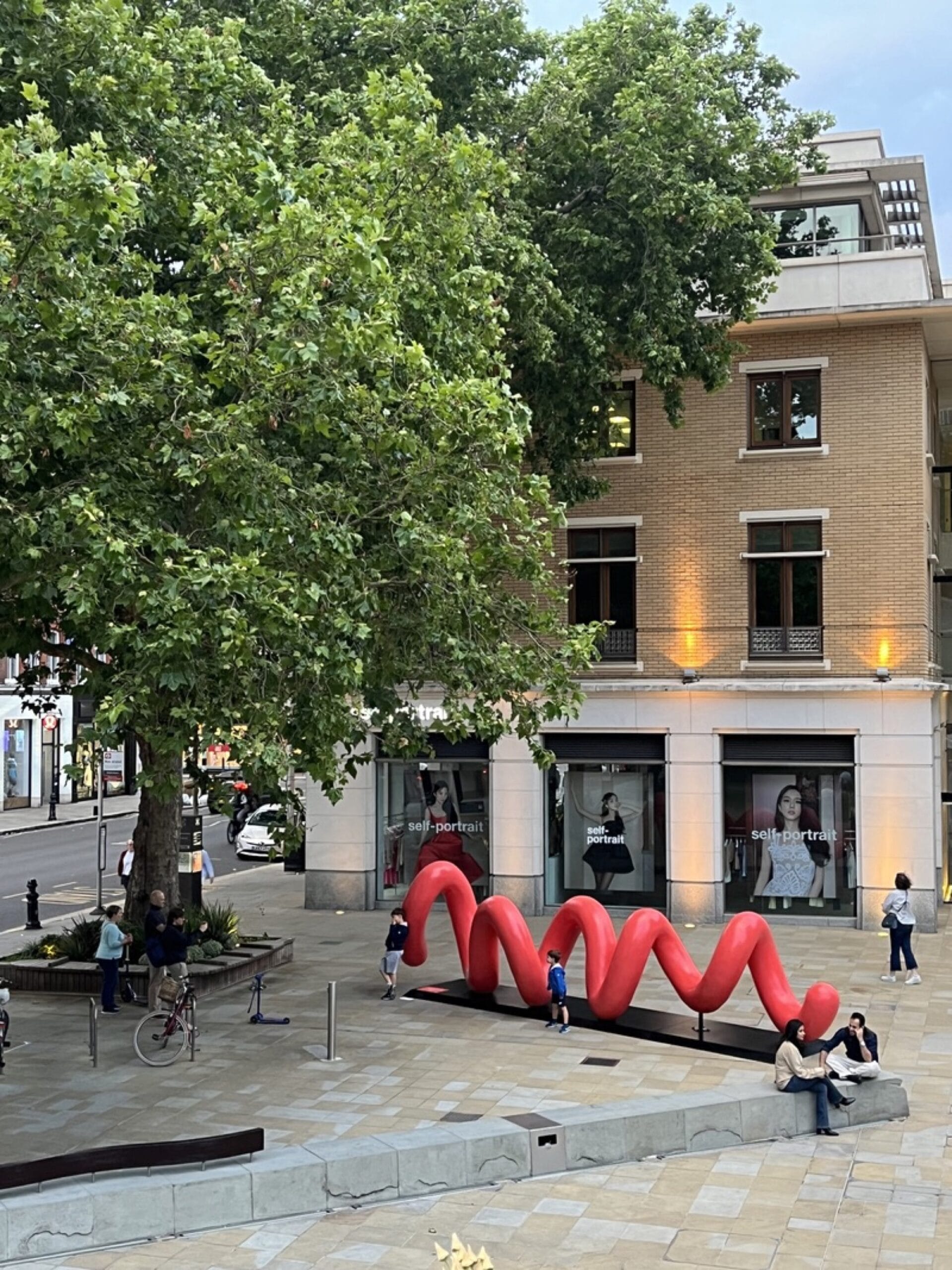 a large-scale, red public sculpture of curving lines that spell the word "love" when viewed from the right perspective, shown in this image from a high vantage point in London's Duke of York square