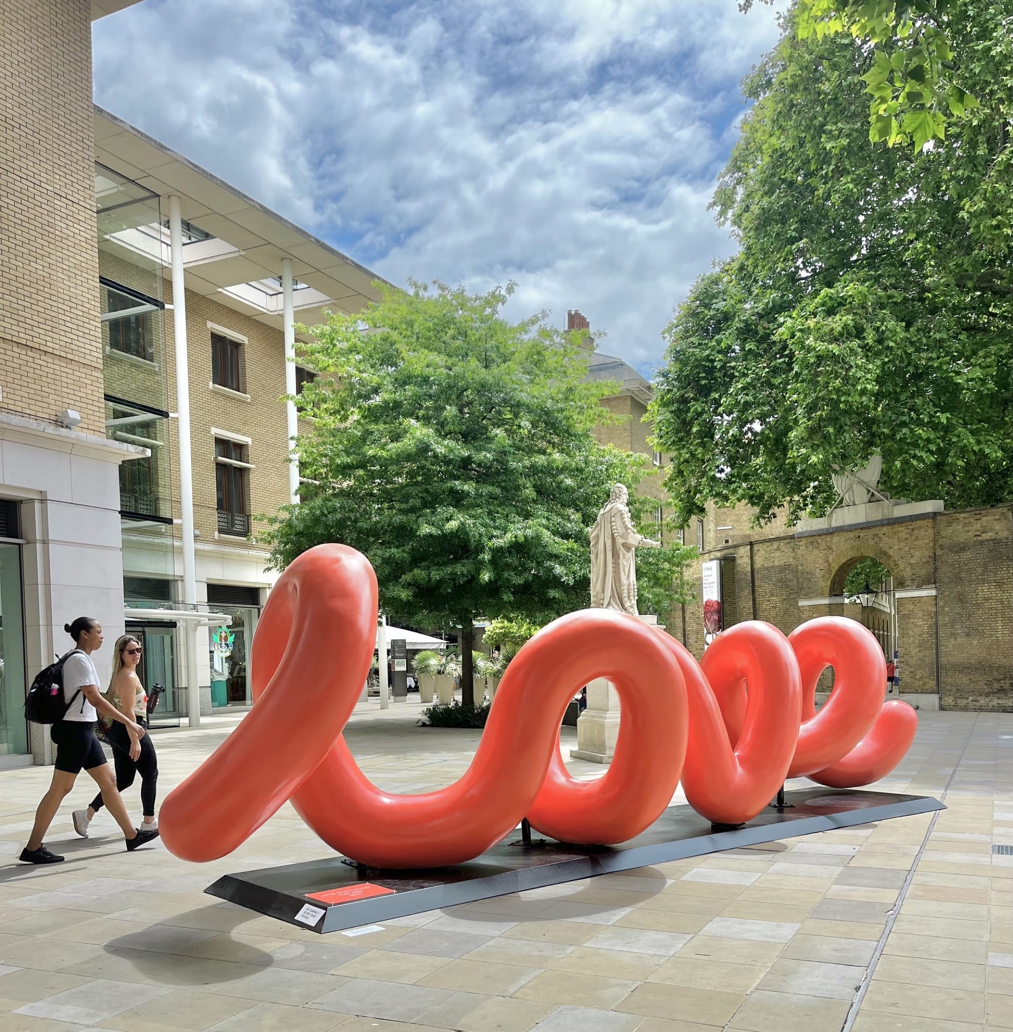 a large-scale, red public sculpture of curving lines that spell the word "love" when viewed from the right perspective, shown in this image with two people walking past it in Duke of York Square in London